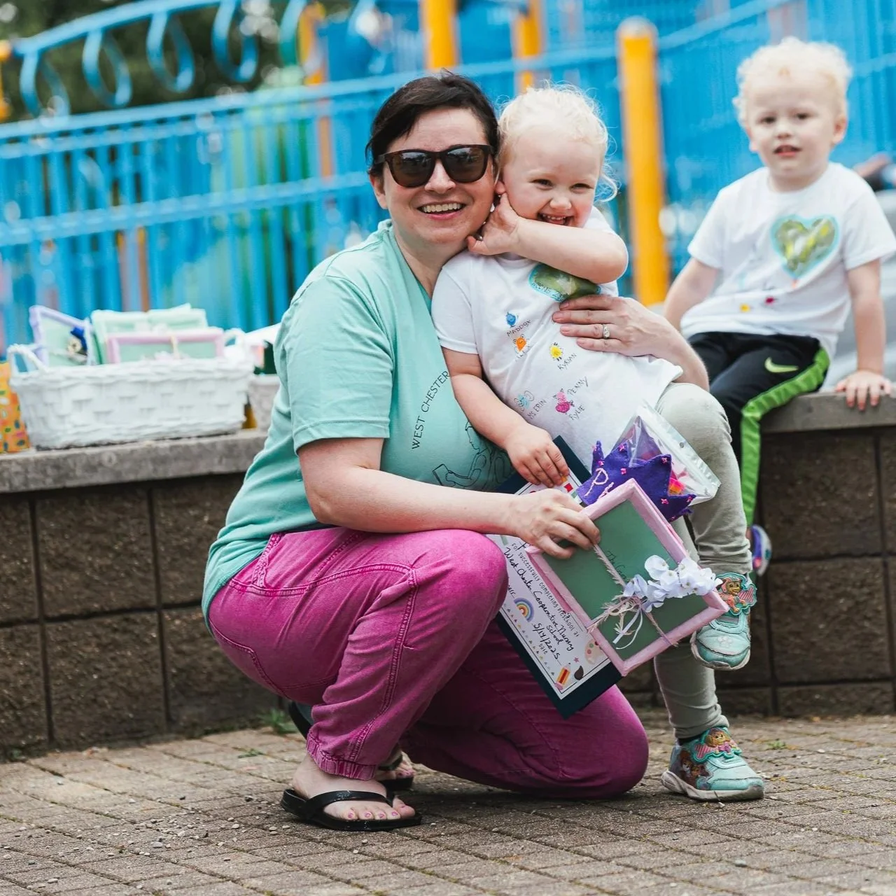Woman in sunglasses and pink pants kneeling on the ground, smiling and holding children, one child hugging her neck. The woman holds a certificate and a small gift box, with two young children in casual clothes nearby. They are outdoors, with a colorful playground fence in the background.