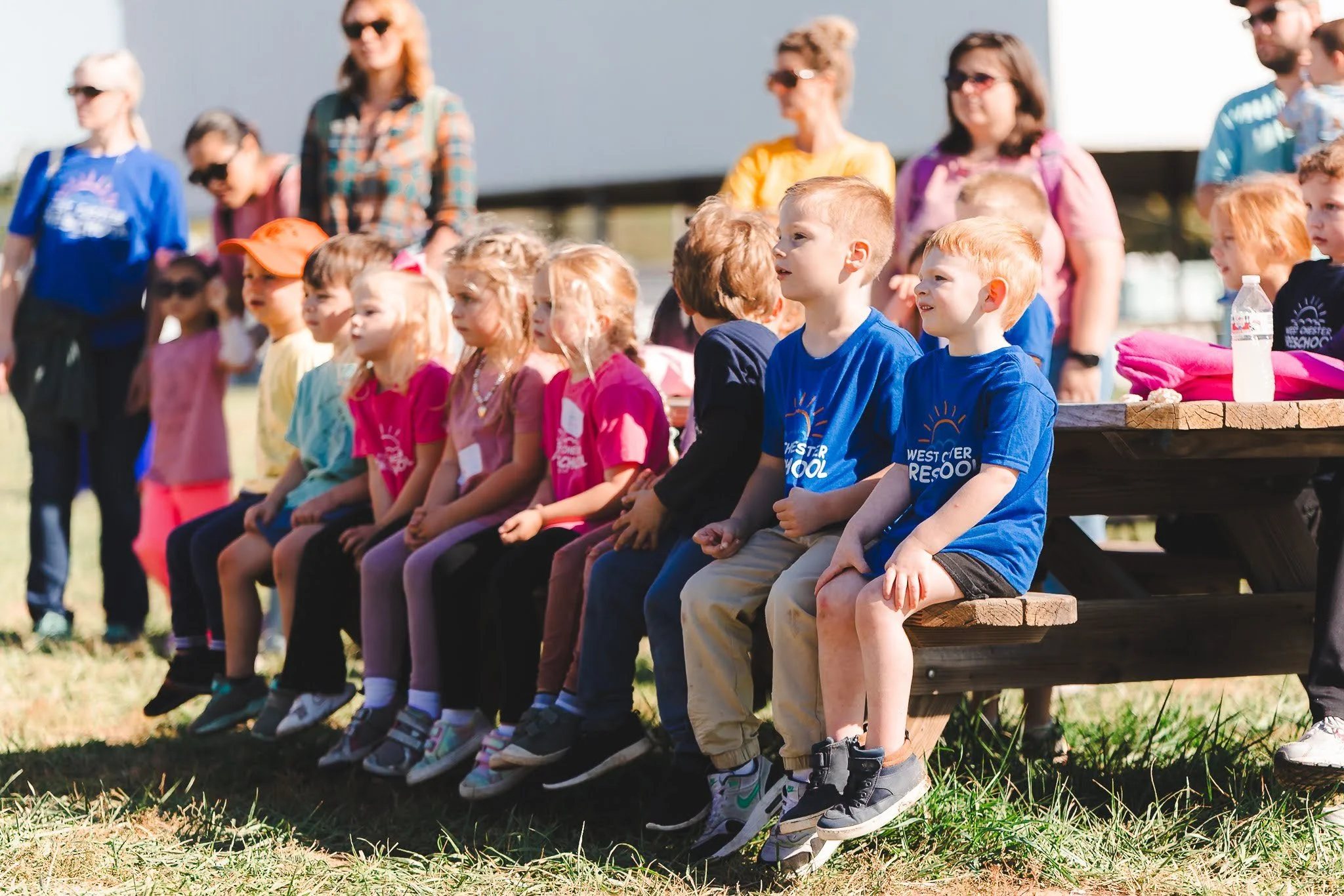 Children sitting on a wooden bench outdoors, some wearing colorful shirts, with adults standing behind them. The children appear to be at a school or community event.