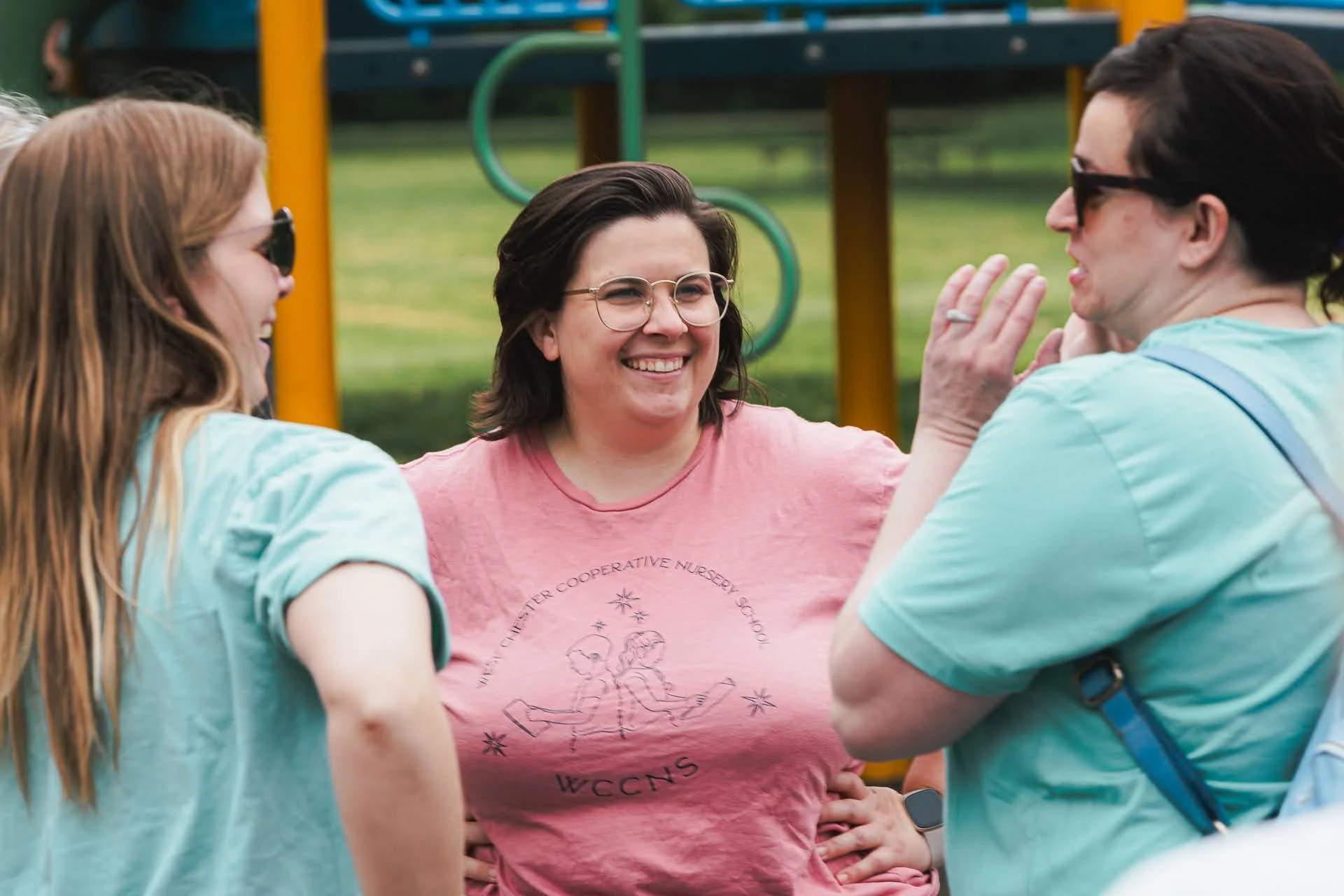 Four women are smiling and conversing outdoors in front of a playground. The woman in the center is wearing glasses and a pink T-shirt with a graphic of two horses and text. The other women wear sunglasses and light-colored shirts.