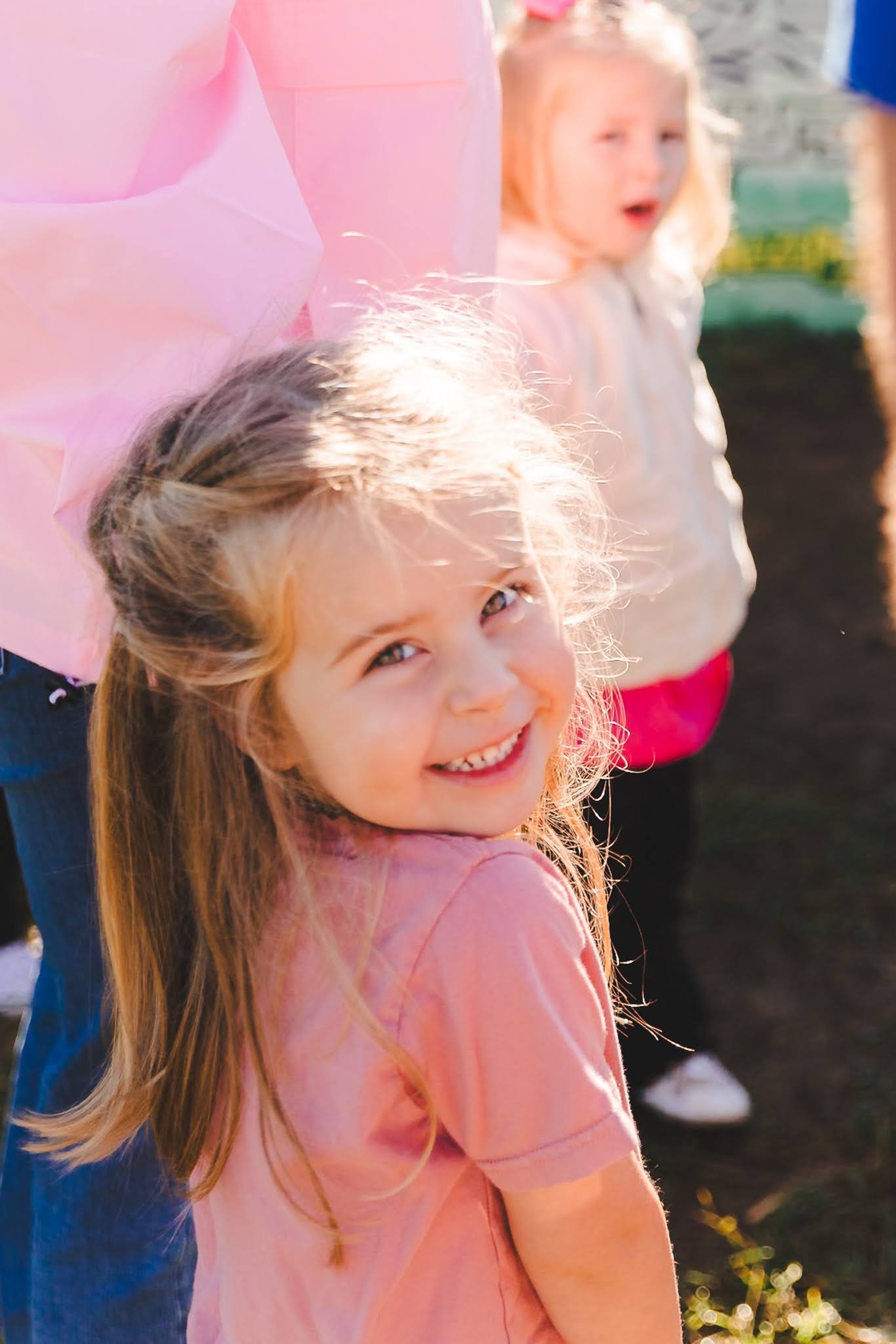 Young girl with blonde hair smiling at the camera, wearing a pink shirt, outdoors with two other children in the background.