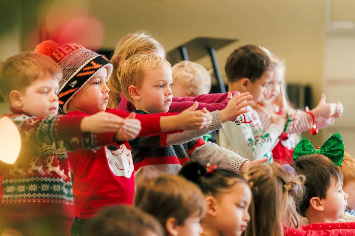 Children dressed in holiday sweaters and accessories, sitting and standing in a classroom, participating in a Christmas or holiday activity.