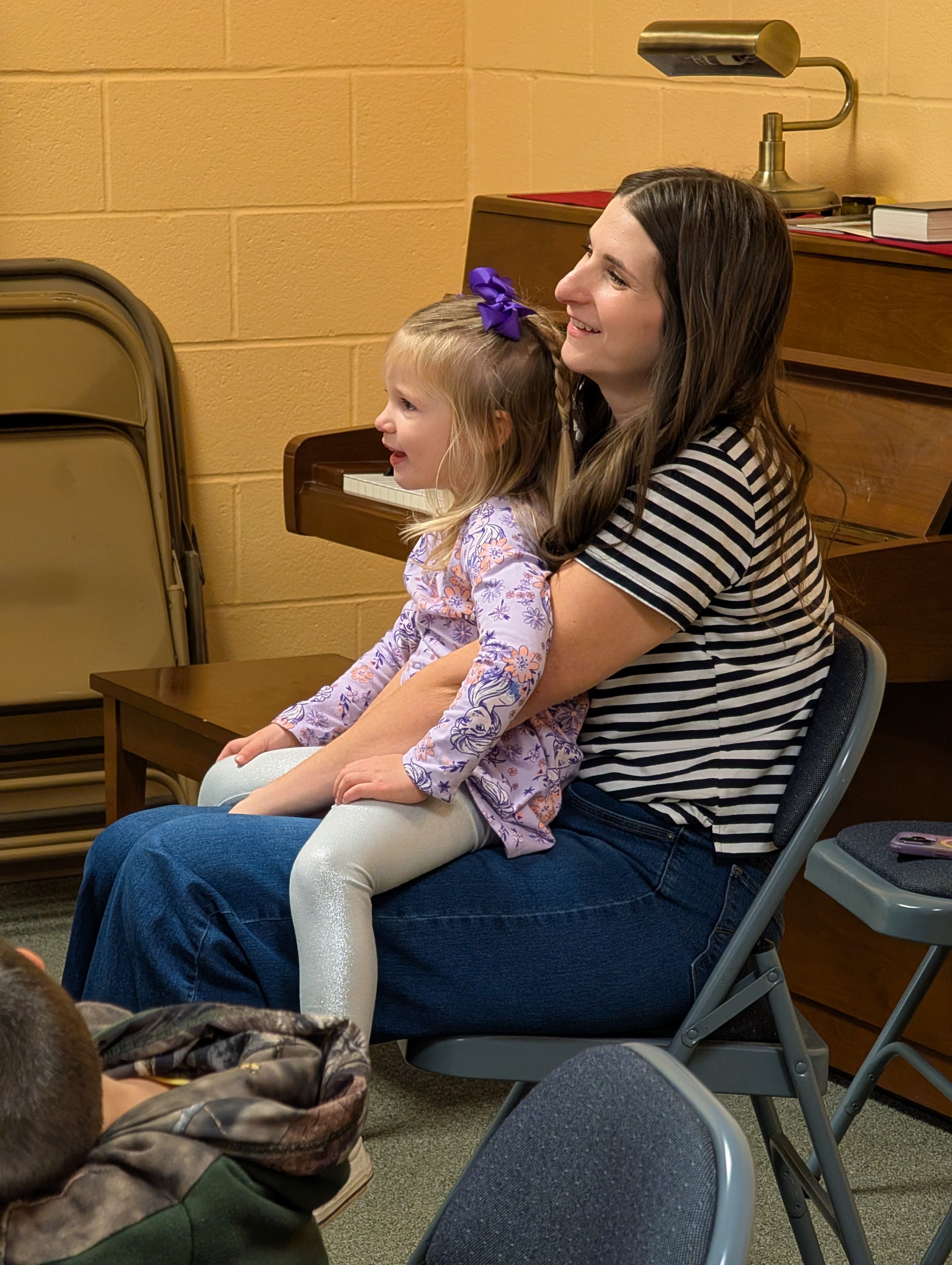 A woman and a young girl sitting on a chair, smiling and watching something in front of them in a room with yellow brick wall and furniture.