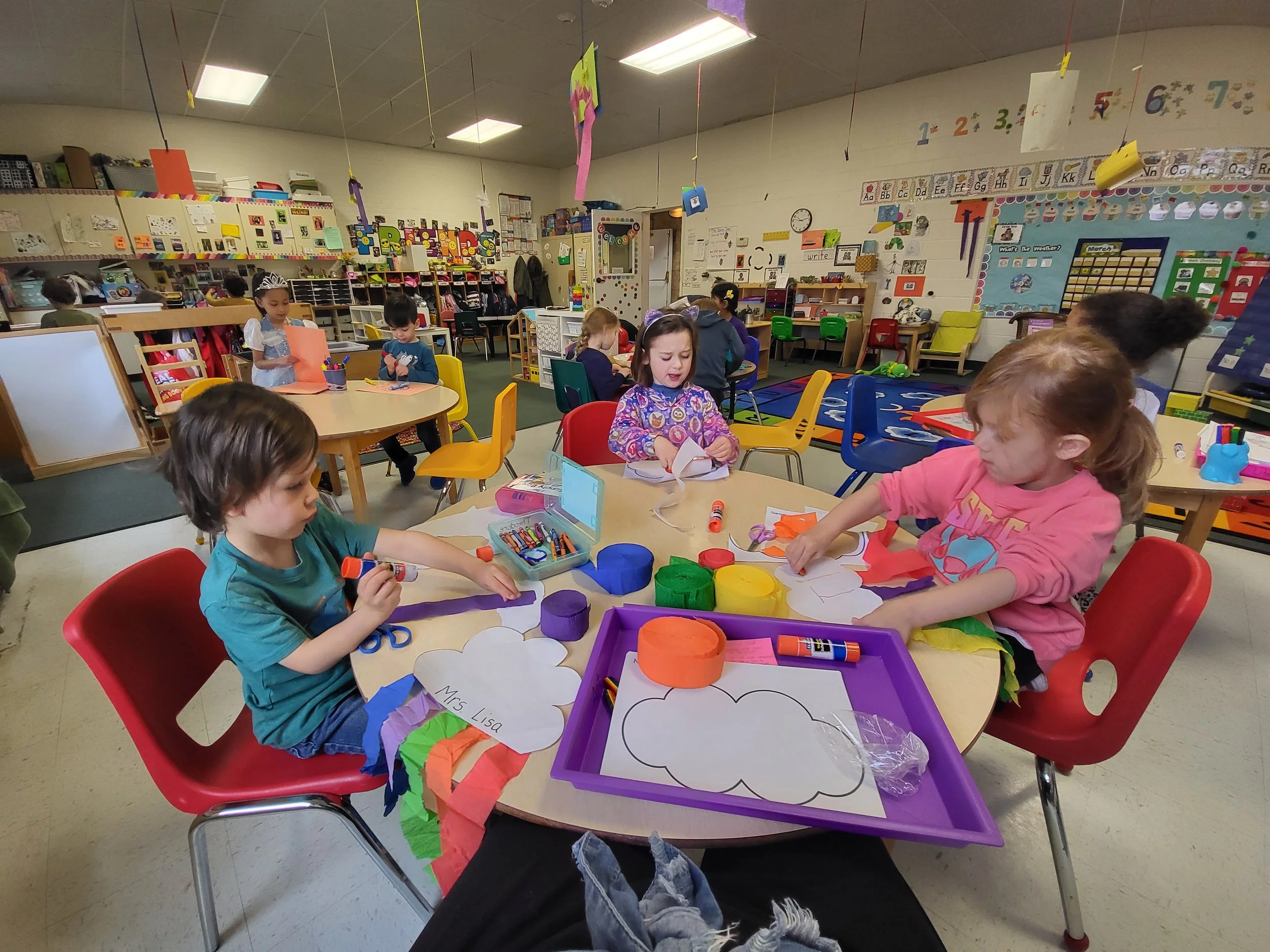 Children engaged in arts and crafts activities at a classroom table, with colorful supplies and paper clouds, in a lively preschool classroom decorated with educational posters and colorful decorations.