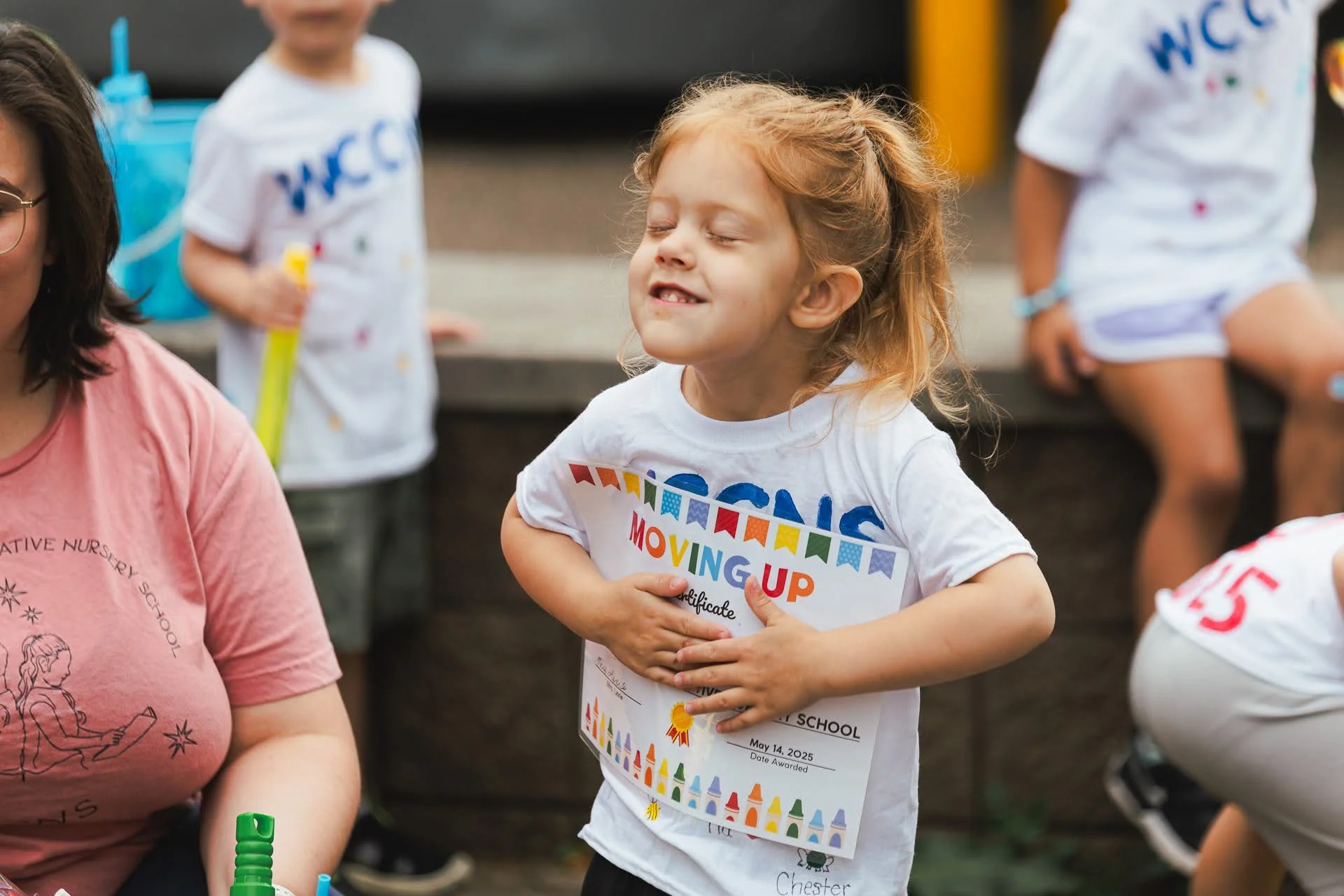 A young girl with red hair is smiling and holding her stomach, celebrating a moving-up ceremony at school. She's wearing a white T-shirt with colorful printing and has her eyes closed. Other children are around her, some sitting on steps, wearing T-s