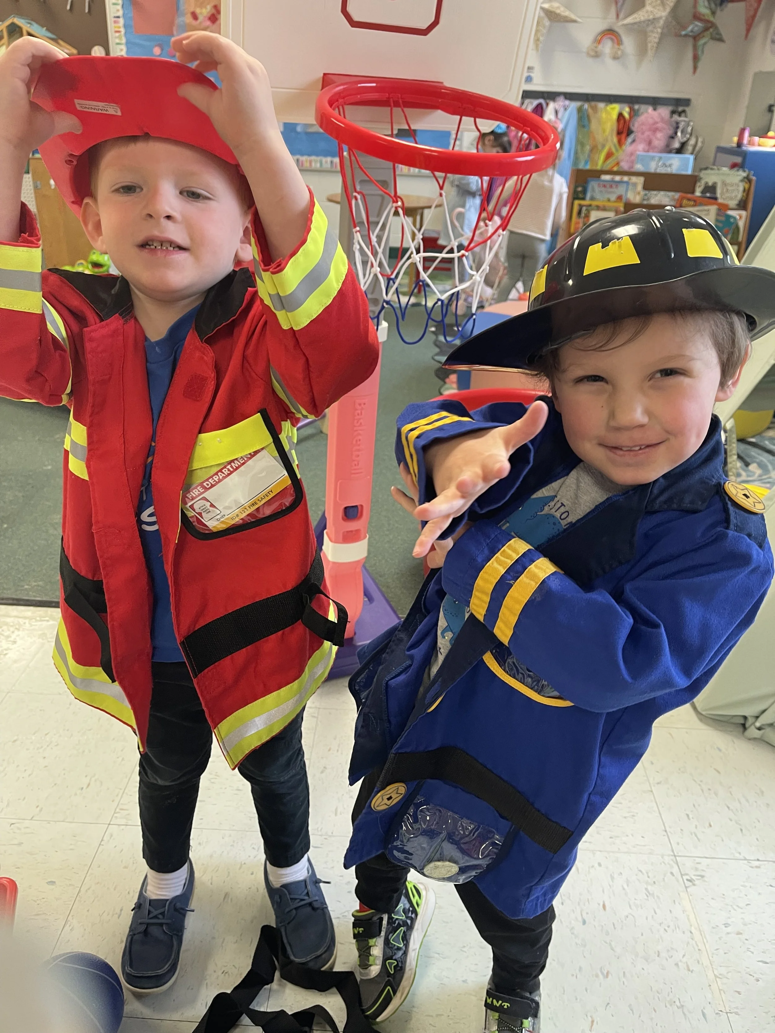 Two young children dressed as firefighters, wearing firefighter costumes with hats, standing indoors next to a small basketball hoop. One child appears to be adjusting a firefighter helmet, and the other is holding something above his head.