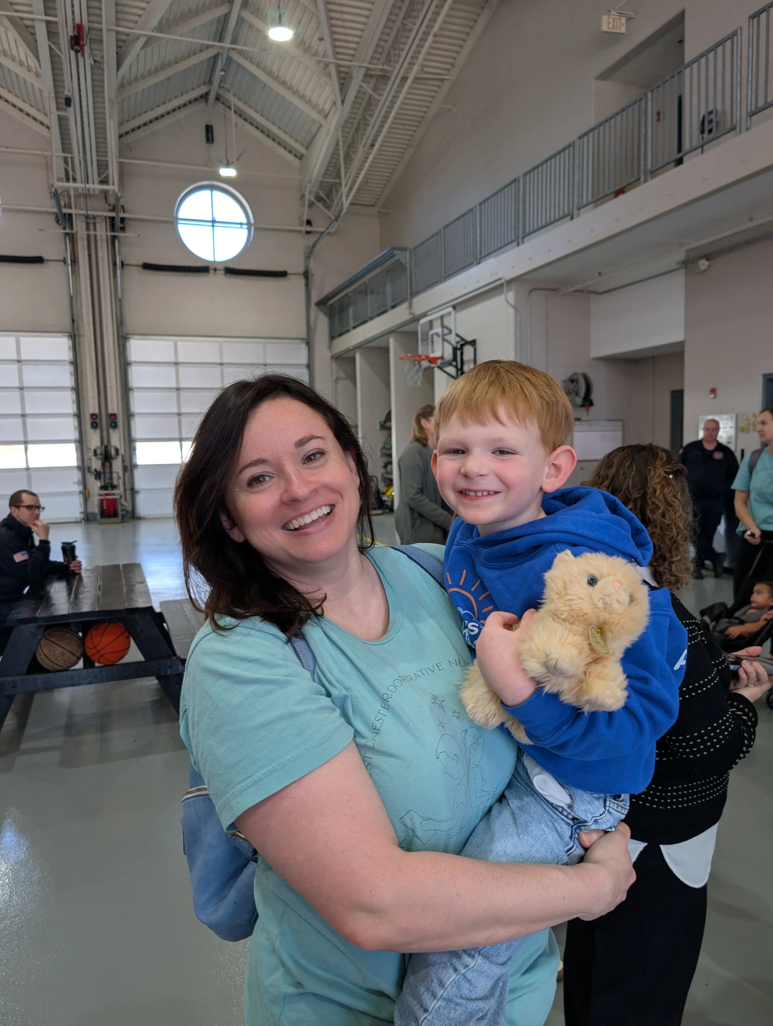 A woman holding a young boy in a large indoor space, smiling at the camera. The boy is holding a plush teddy bear and wearing a blue hoodie. There are other people in the background near a basketball hoop, and the space appears to be a gym or recreat