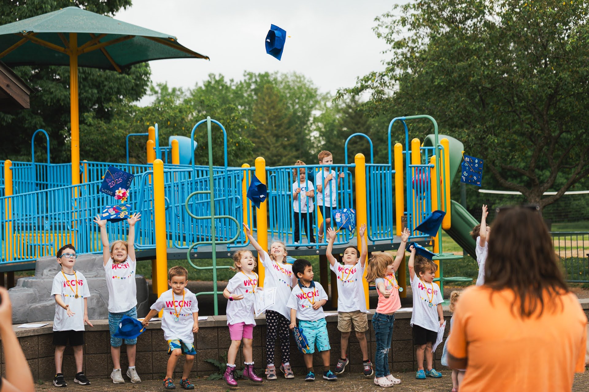 Children in wccns t-shirts celebrating graduation on a playground with a slide, with their mortarboards thrown in the air and an adult supervising.