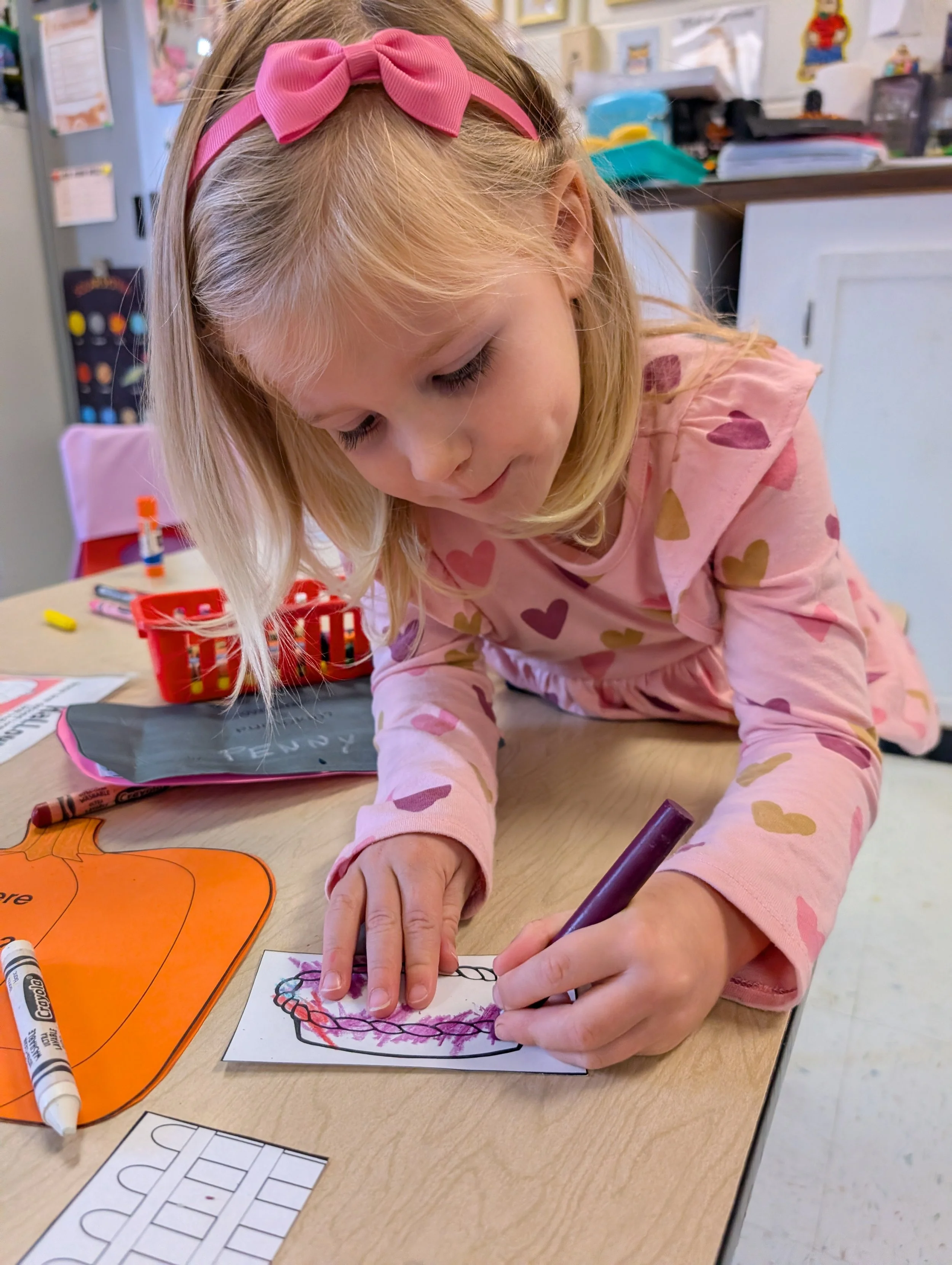 A young girl with blonde hair and a pink bow headband coloring a pumpkin picture on paper at a table with crayons and craft supplies in the background.