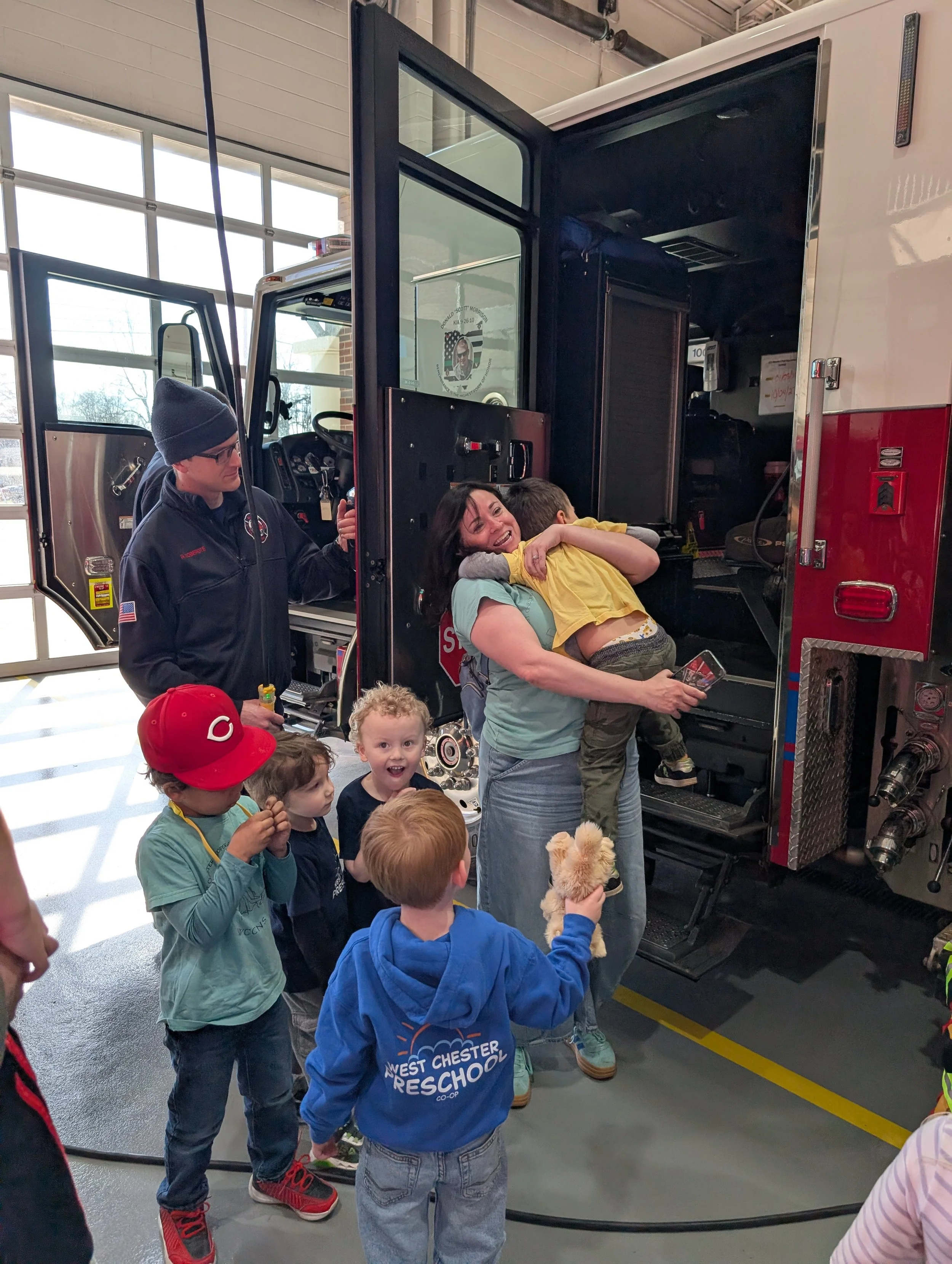 A group of children and a woman hugging a firefighter inside a fire station, near a fire truck.