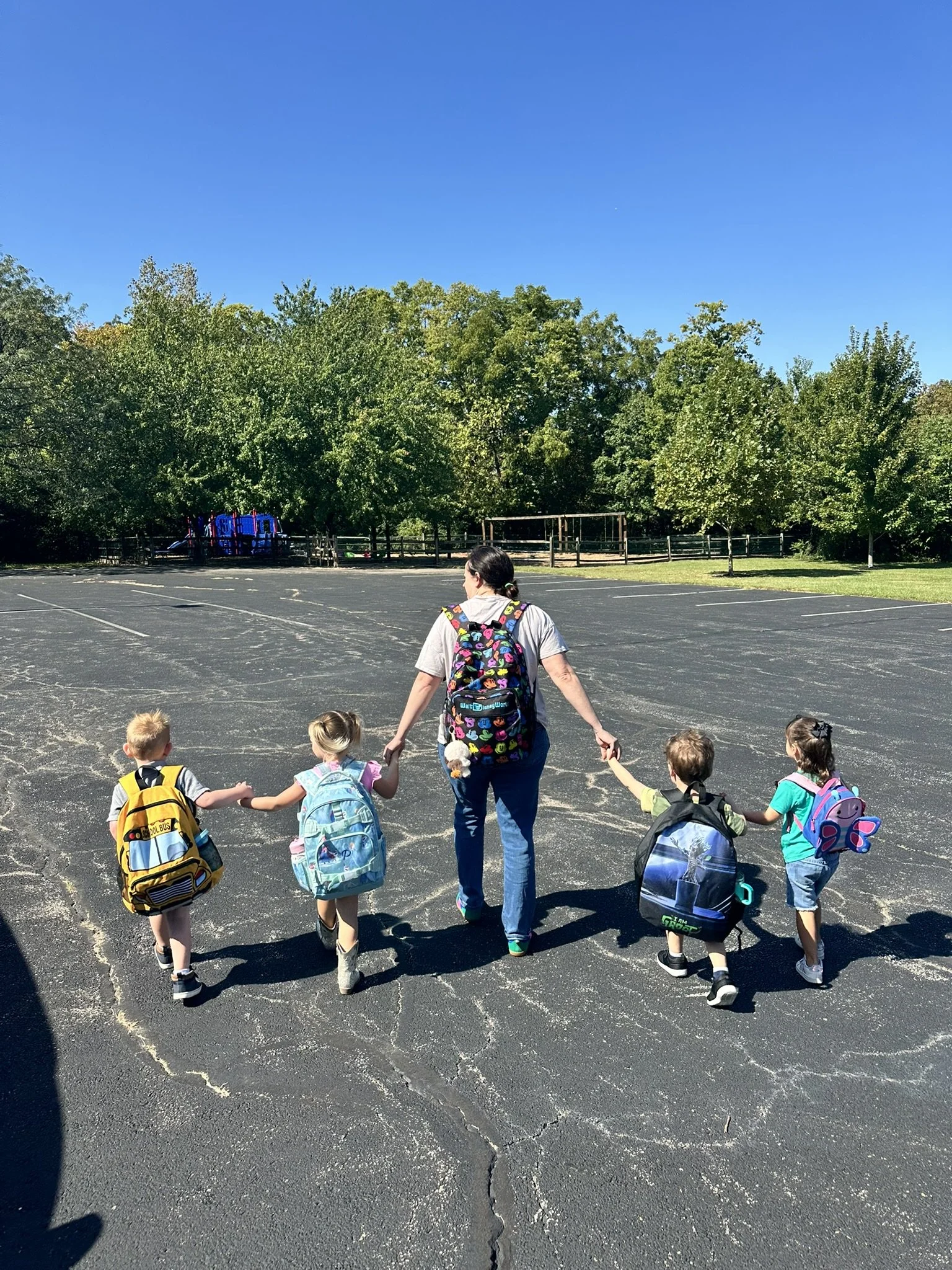 A woman holding hands with four young children as they walk across an empty parking lot on a sunny day, with trees and playground equipment in the background.