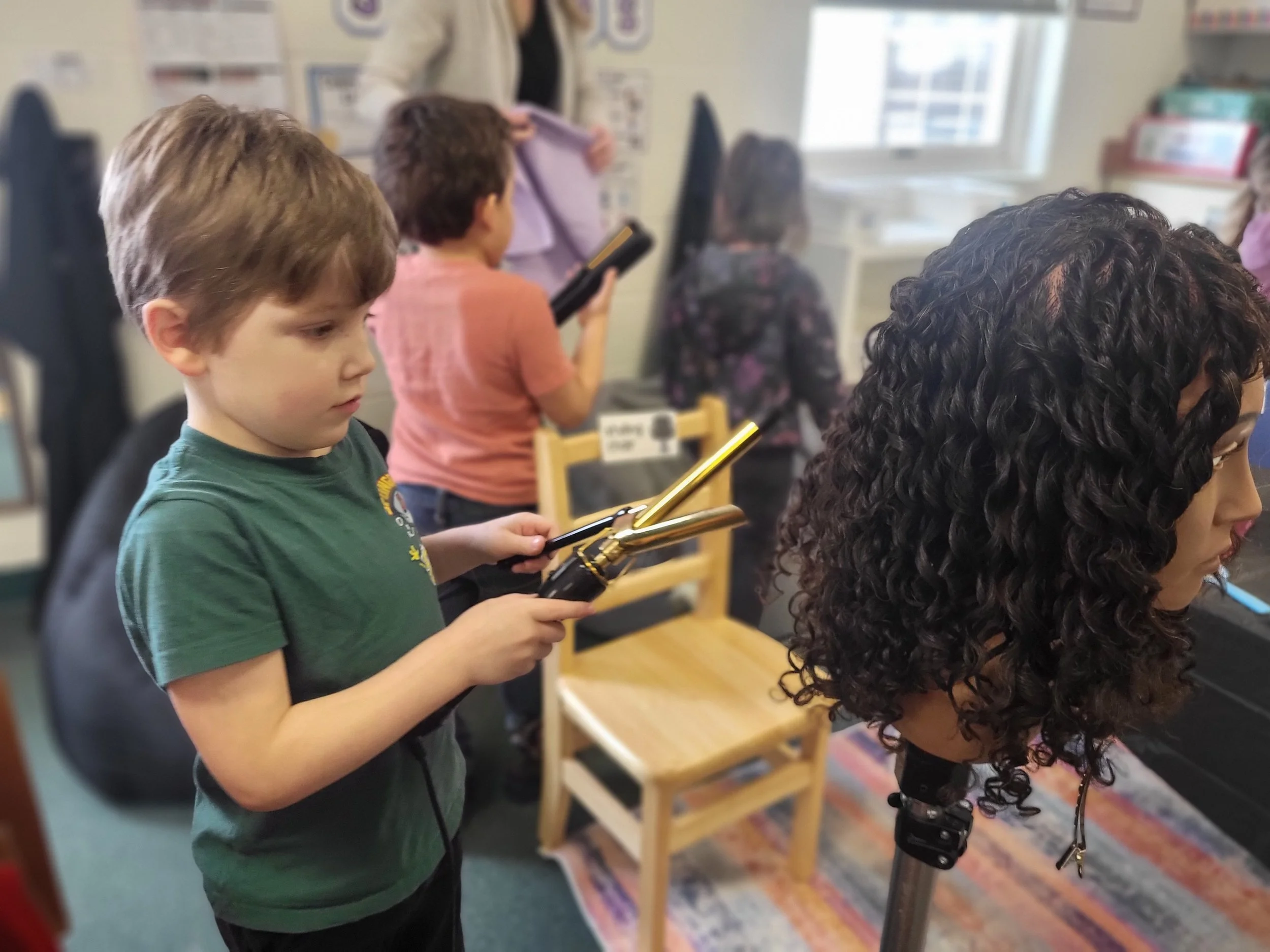 Children in a classroom holding hair styling tools, standing near a mannequin head with curly hair.