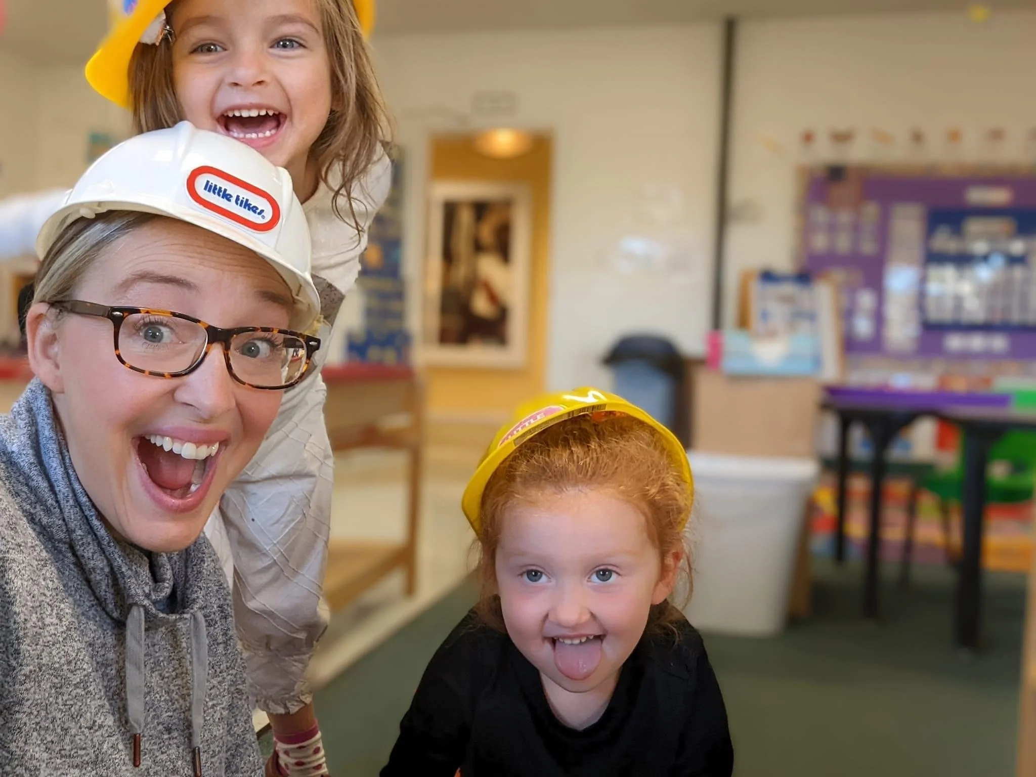 Three children and an adult in a classroom wearing yellow and white safety helmets, all smiling and making silly faces.