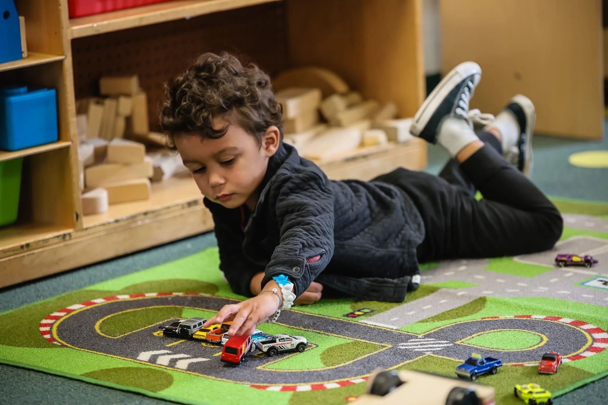 A young boy lying on his stomach on a colorful play mat with a race track design, playing with toy cars and trucks.