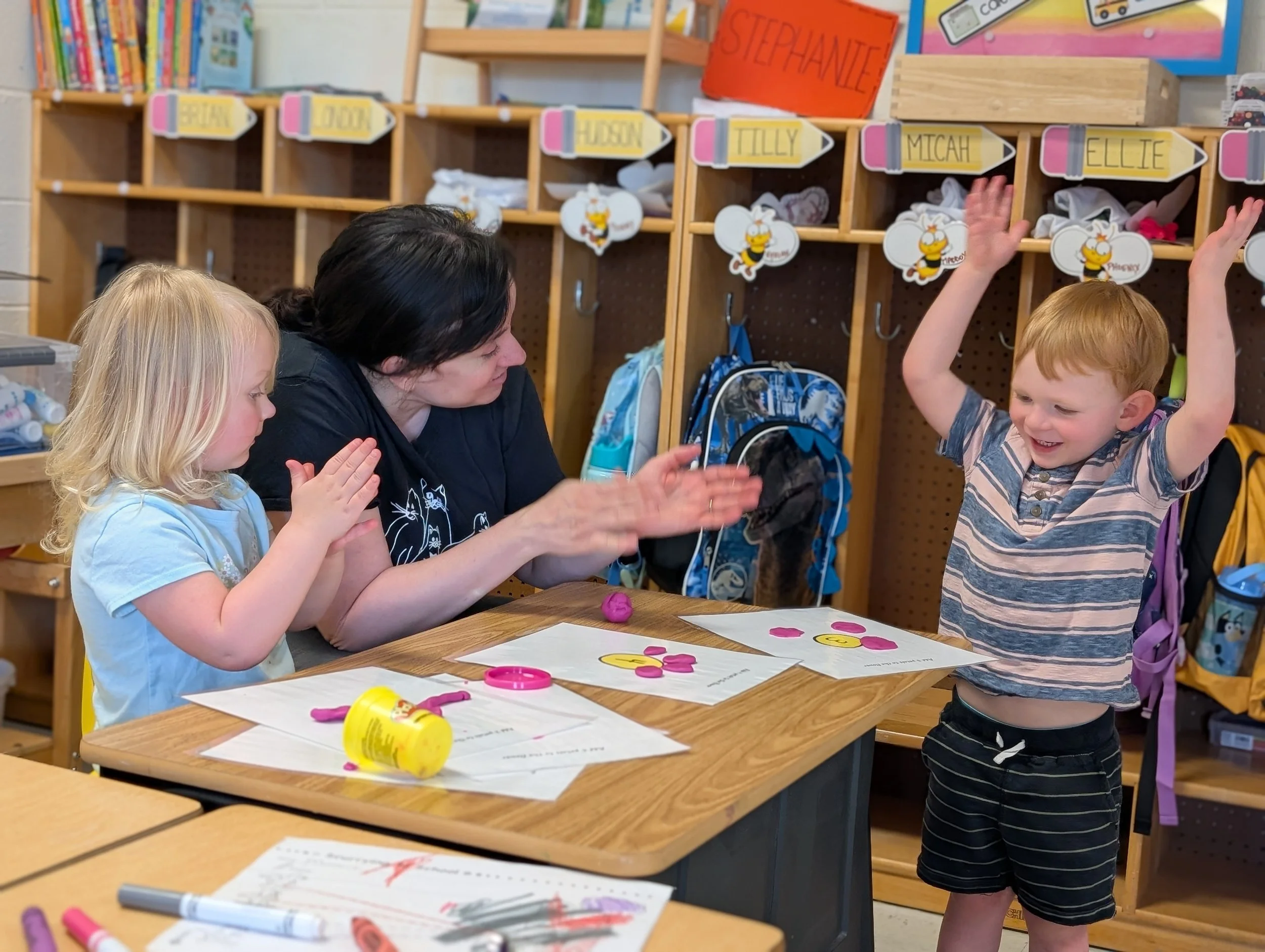 A young boy with red hair and a striped shirt is smiling with his arms raised, celebrating, while sitting at a table with a woman and a girl in a classroom. The woman and girl are clapping and watching him. There are papers, markers, and Play-Doh on 