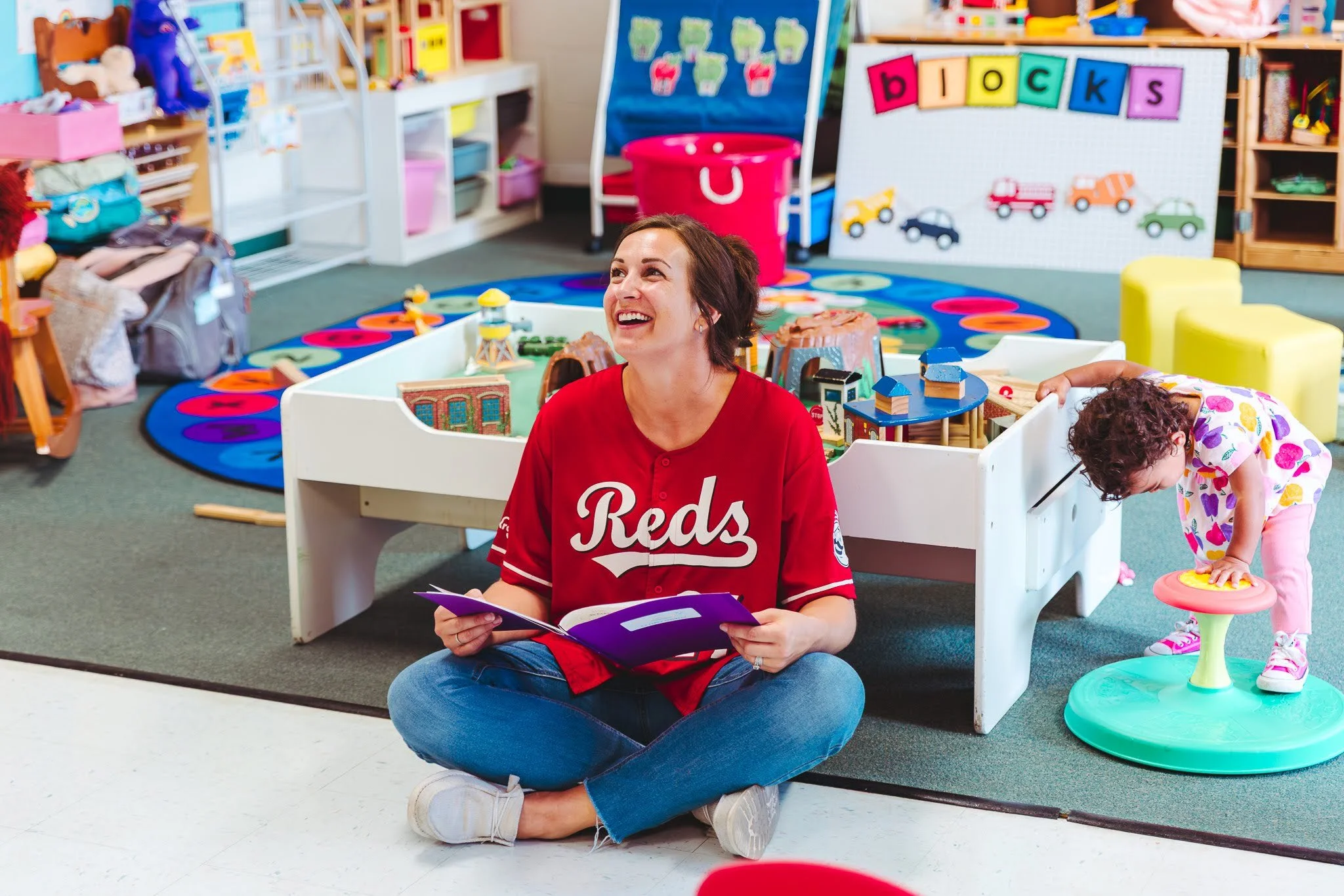 A woman sitting cross-legged on the floor reading a book inside a colorful preschool or daycare classroom. There is a young girl playing nearby with a toy.