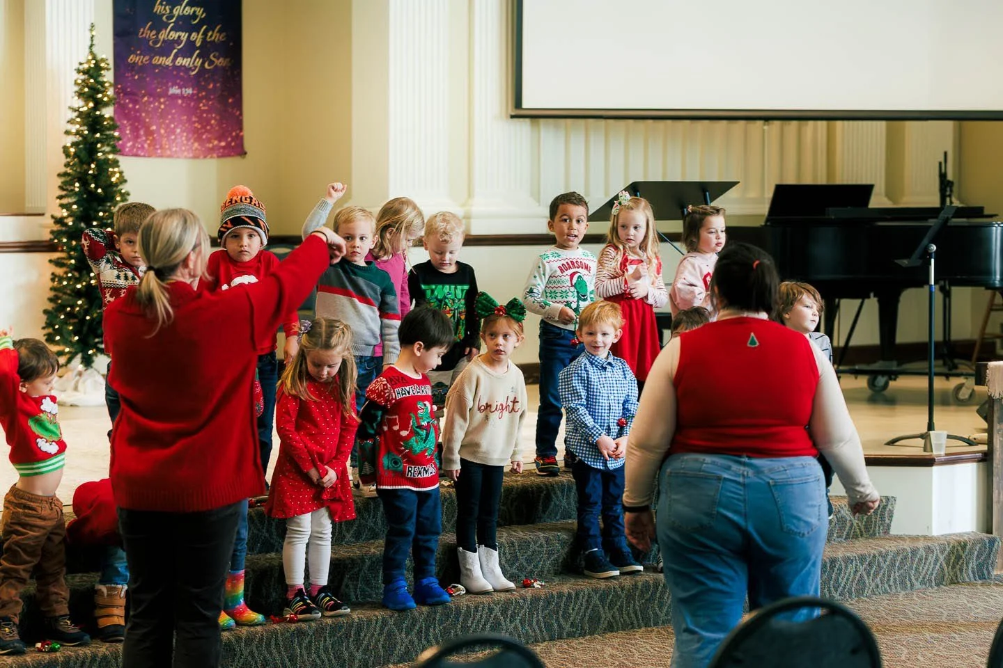 Children on stage singing Christmas carols, with two adults leading, in a decorated room with a Christmas tree and a purple banner.