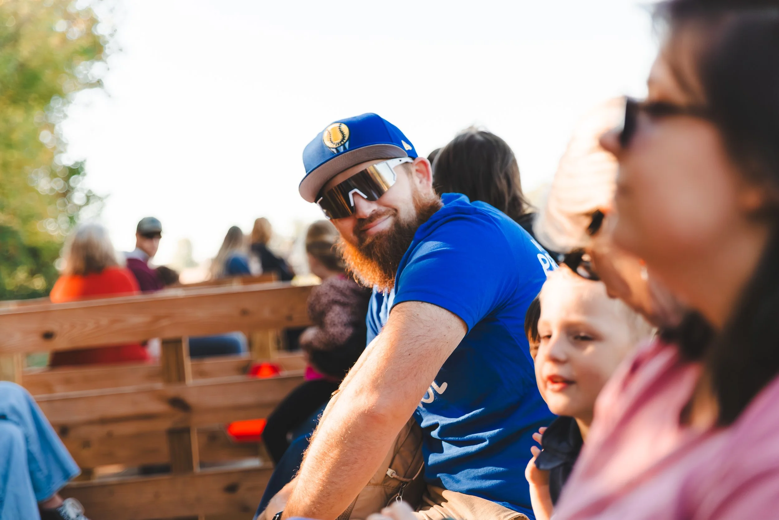 A man with a beard wearing a blue shirt, sunglasses, and a baseball cap, sitting outdoors at a gathering with other people and children, on a sunny day with trees in the background.