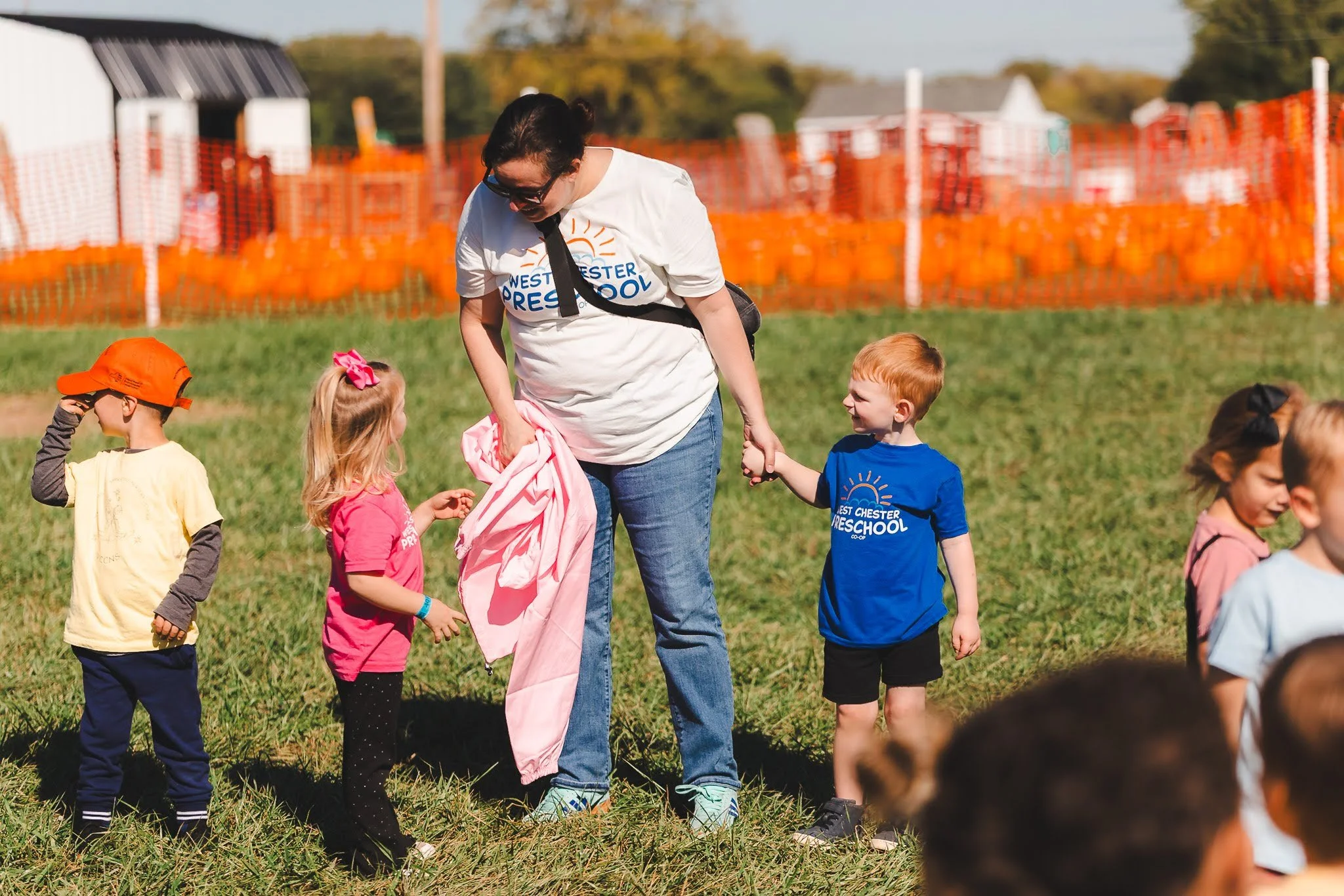 A woman and children at an outdoor preschool event, with children wearing colorful clothes, some holding hands, and orange pumpkin decorations in the background.