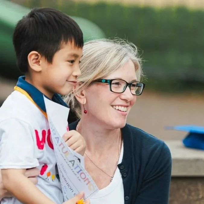 A young boy and a woman with glasses smiling outdoors; the boy is holding a certificate, and the woman is wearing a navy cardigan.