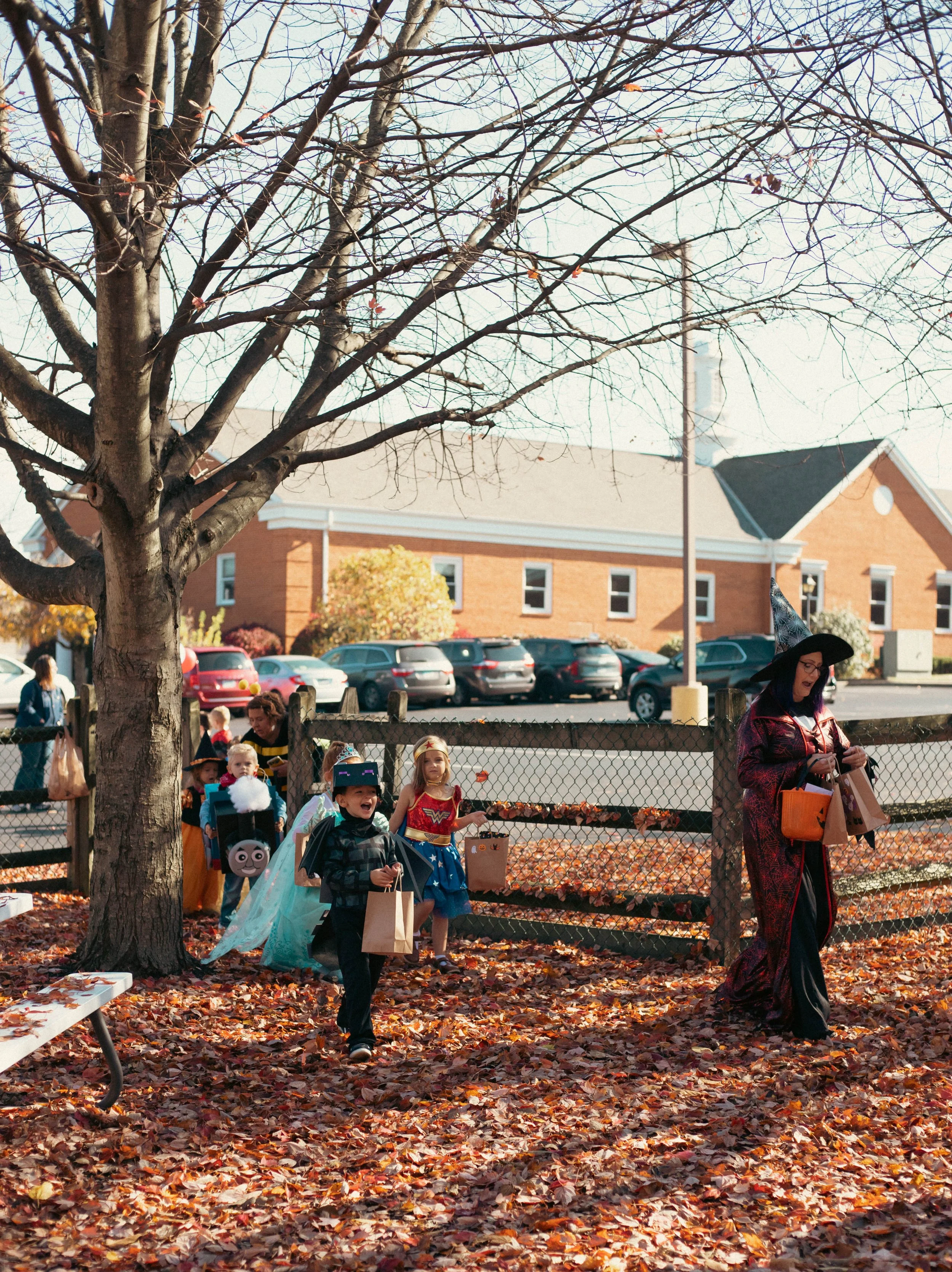 Children dressed in costumes for Halloween trick-or-treating, walking along a leaf-covered area near a fenced yard, with an adult woman in costume holding bags, under a large tree with bare branches.