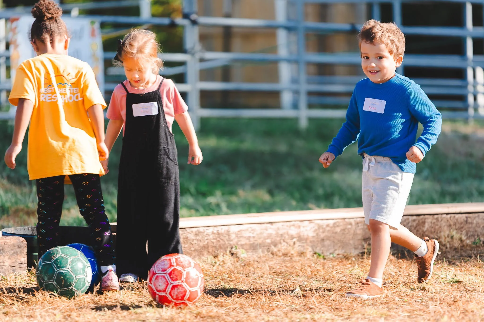 Three young children playing with soccer balls outdoors on a sunny day. Two children stand next to each other near two soccer balls, while a third child runs toward them. The children have backpacks and are in a grassy area with a wooden barrier and 