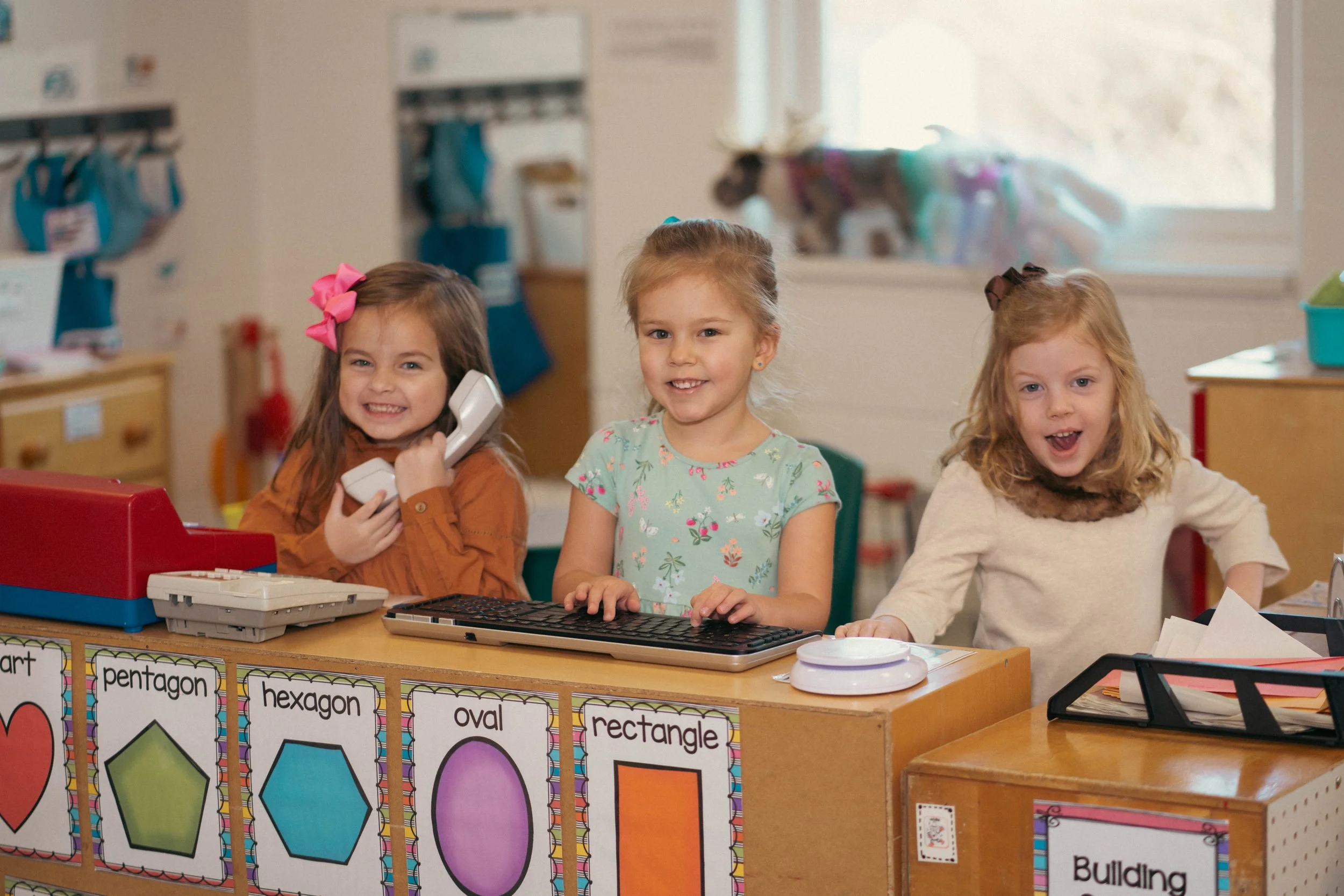 Three young girls in a classroom standing behind a counter with educational posters of geometric shapes and their names.