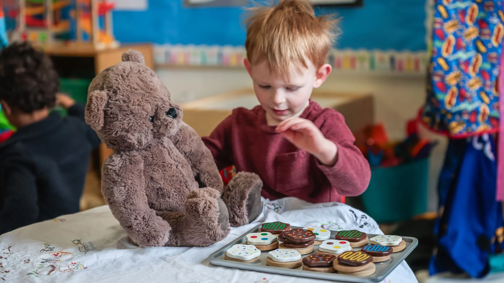 A young boy with red hair in a maroon shirt playing with plush teddy bear and pretend cookies in a classroom or playroom.