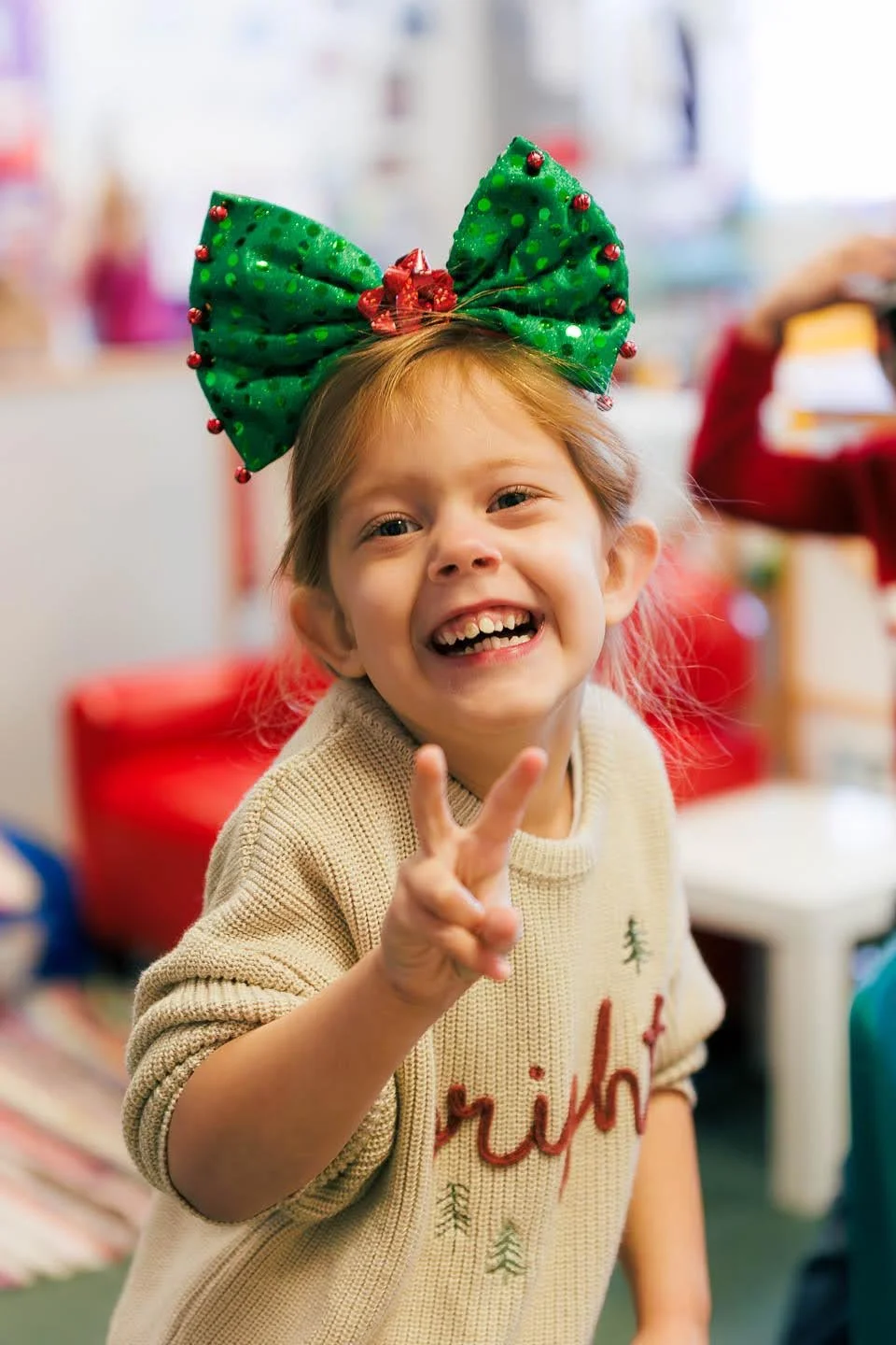 A young girl wearing a large green Christmas bow headband, smiling and making a peace sign, during a festive holiday celebration.