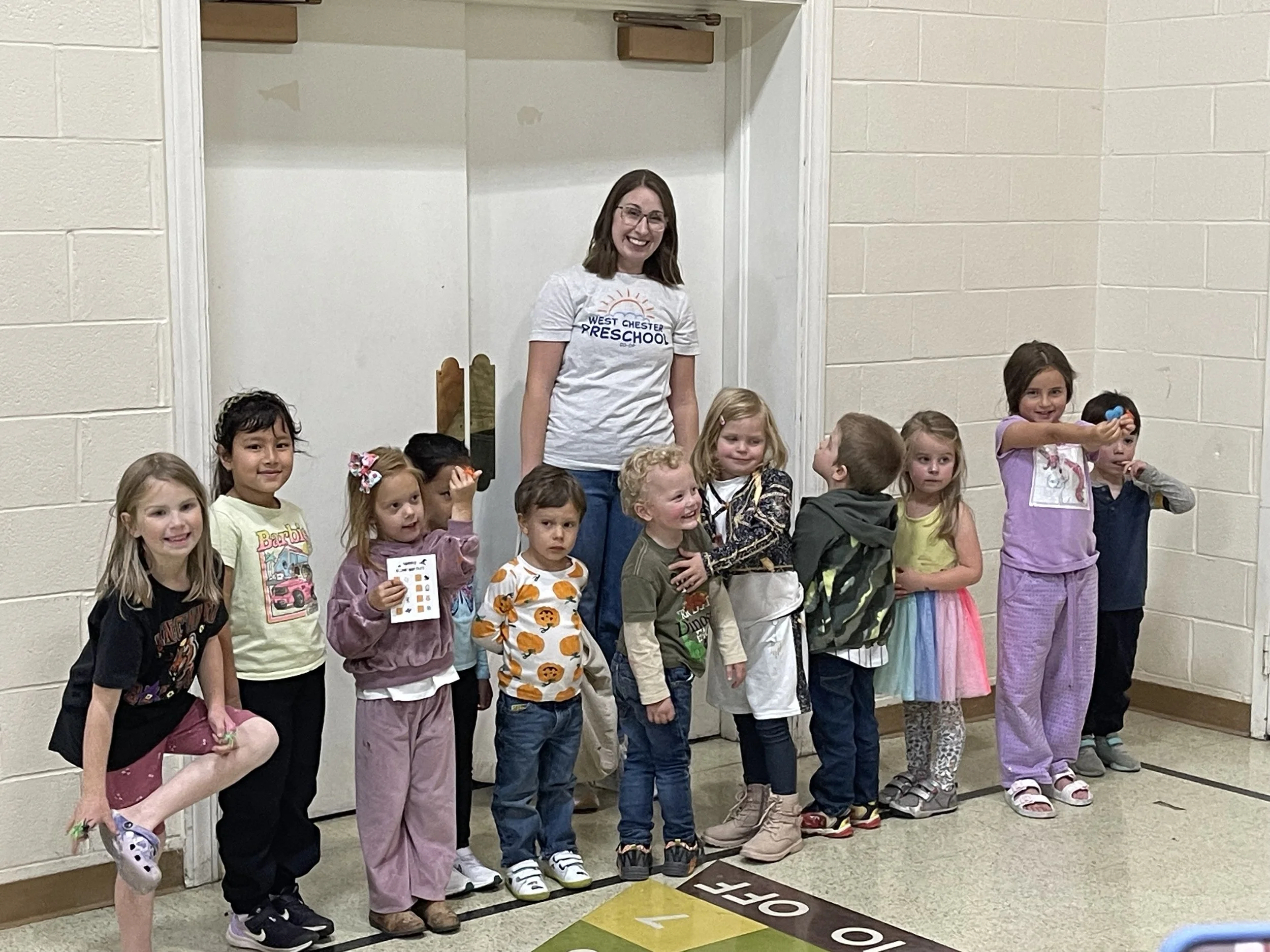 Group of young children with a woman, possibly a preschool teacher, standing in a hallway near a door, with some holding small objects or papers, smiling and posing for the photo.