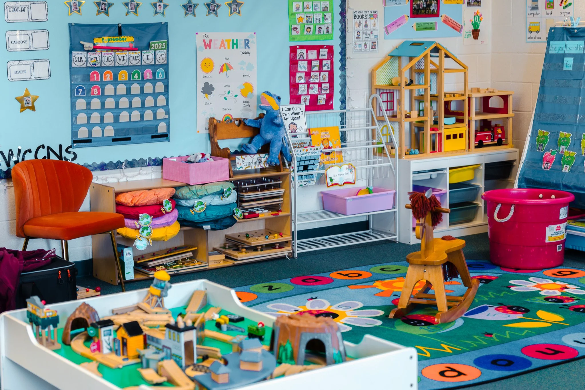 A colorful preschool classroom with educational posters, a moon calendar, weather chart, and various toys. There is a play area with a train set, a rocking horse, and a pink bucket for toys. The room has bright decorations, a rug with numbers and flowers, and shelves filled with books and supplies.
