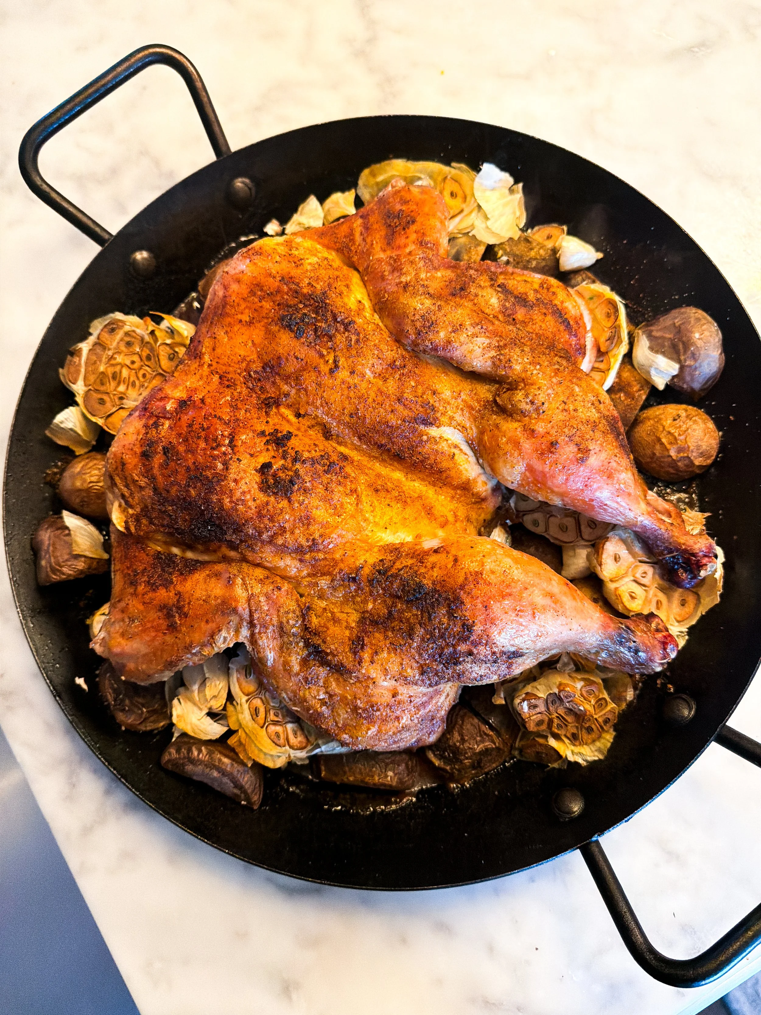 Golden oven-roasted spatchcocked chicken with whole heads of roasted garlic and potatoes in a black paella pan.
