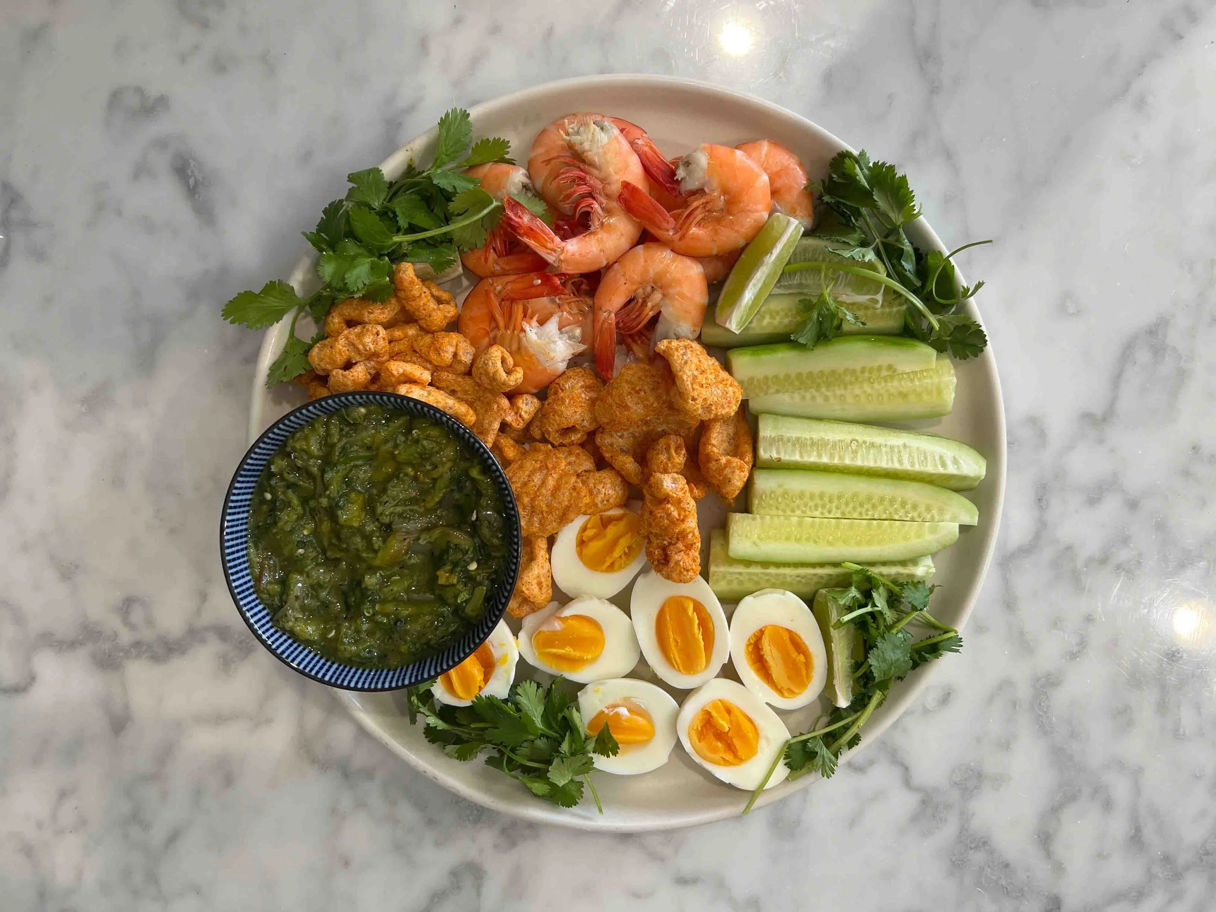 Green nam phrik num in a blue bowl on a platter with pork rinds, halved soft-boiled eggs, cucumber spears, shrimp, and cilantro.