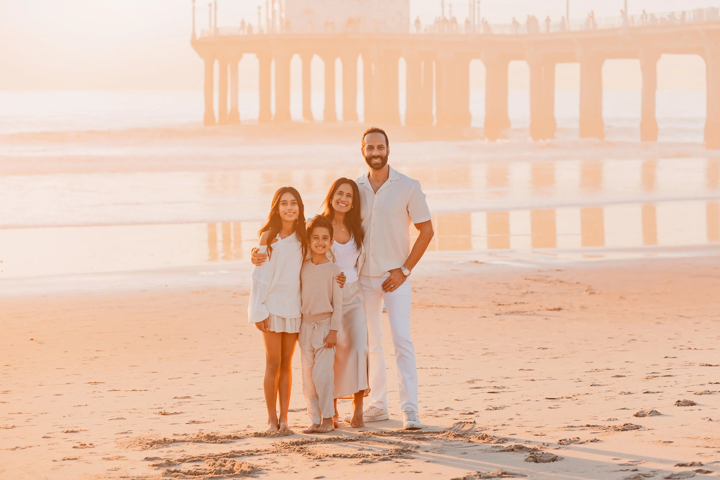 Family of four in white outfits on the beach at sunset with a pier behind them