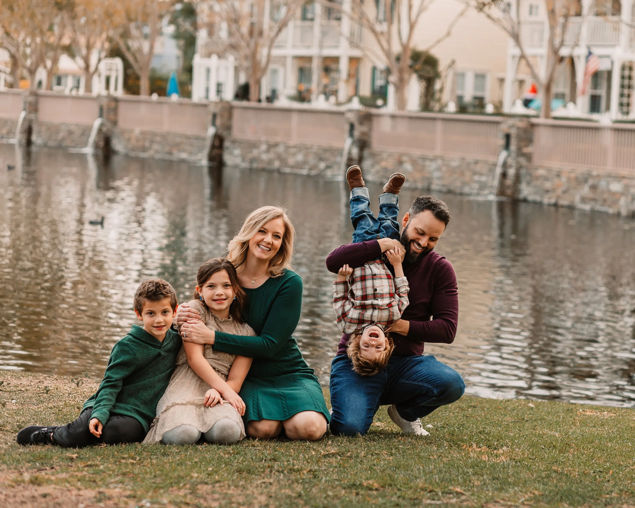 Family of five laughing together by a lake, dad flipping the toddler playfully