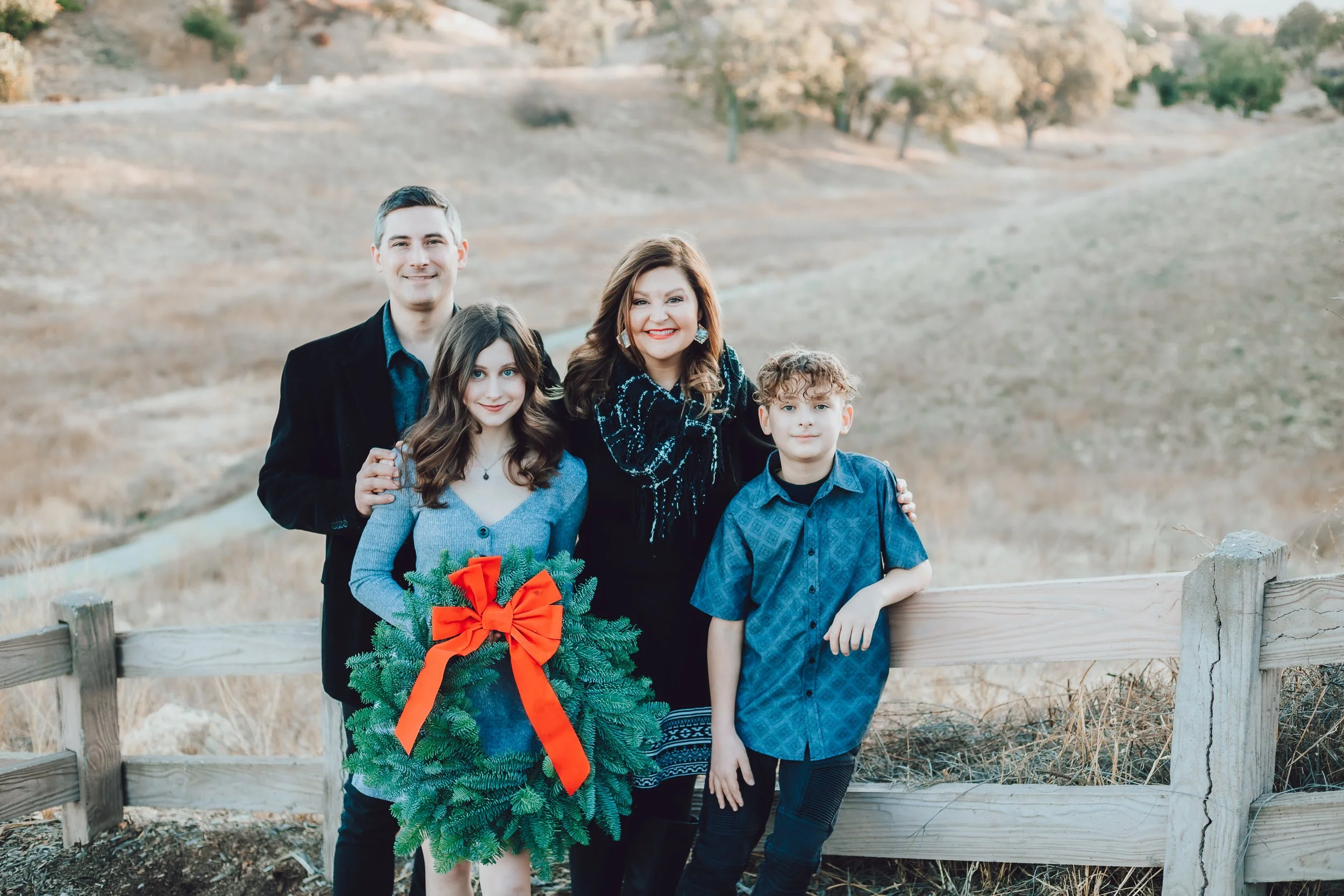 Family of four holding a Christmas wreath with a red bow at a wooden fence in a golden Santa Clarita field at sunset.