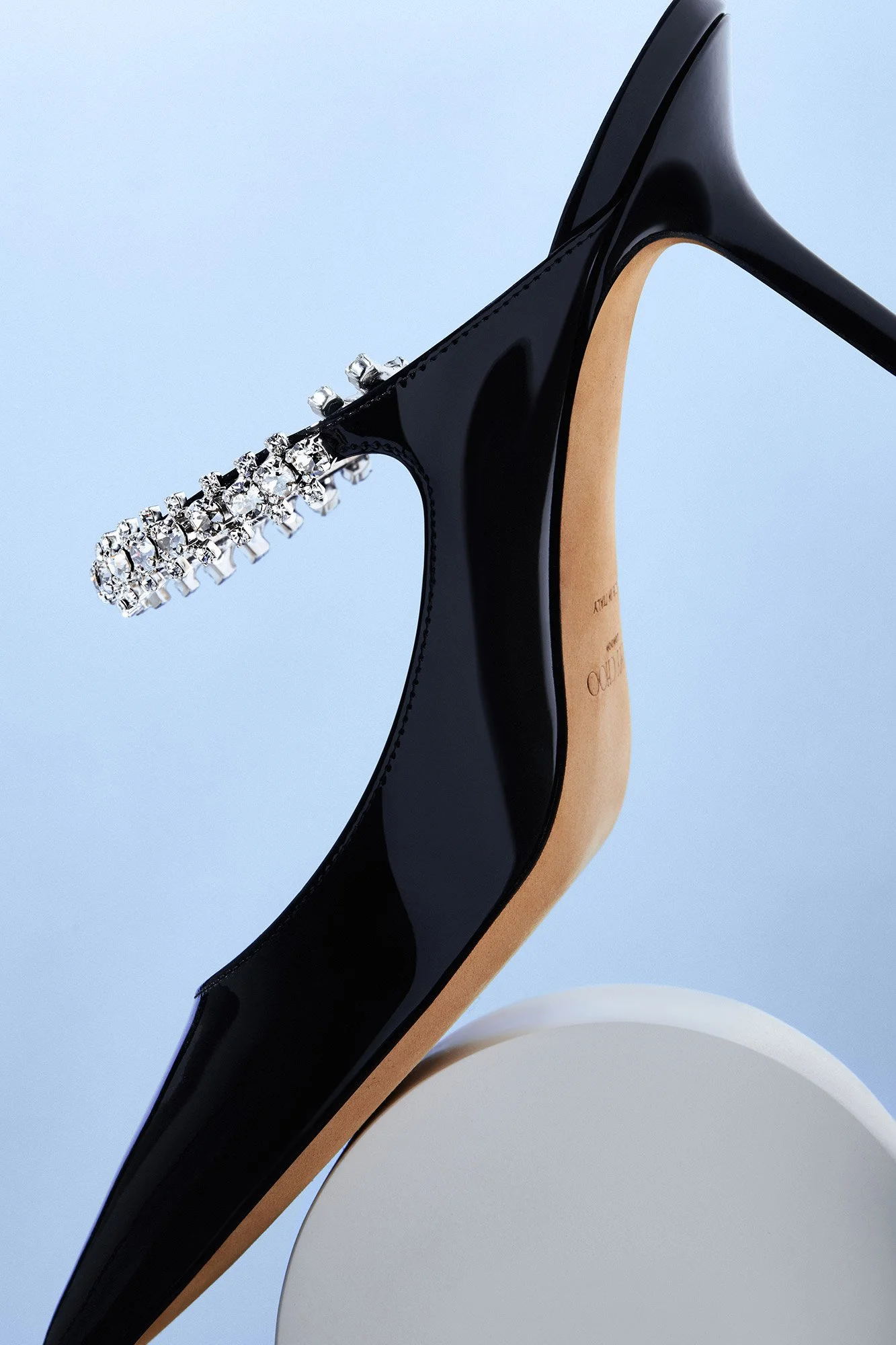 Close-up of a black high-heeled shoe with a decorative strap covered in rhinestones, displayed against a light blue background.