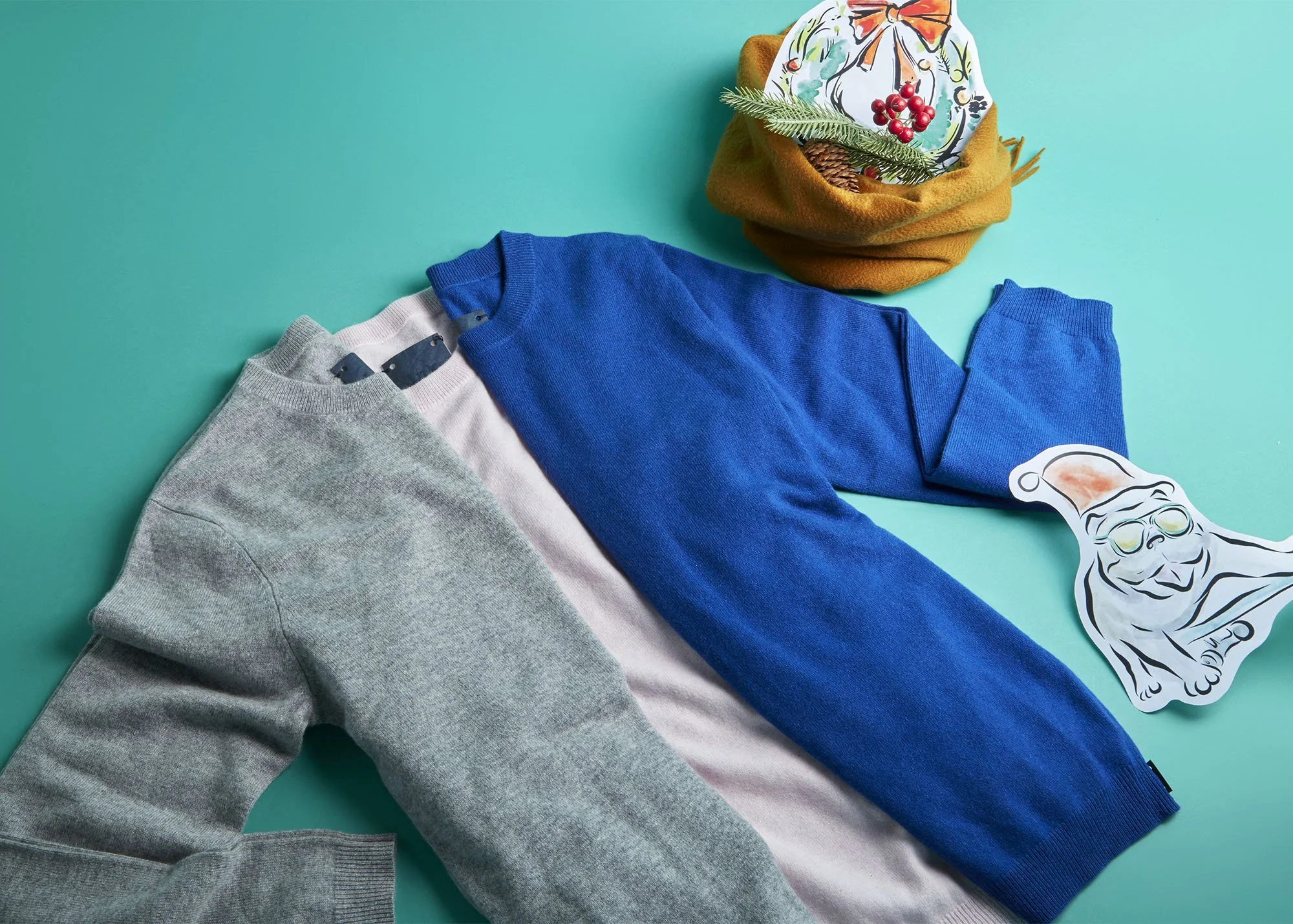 Colorful clothing including a gray sweatshirt, a pink shirt, and a blue sweater laid out on a turquoise surface, with holiday-themed decorations such as a Santa image sticker, a cardboard Santa cutout, and a small Christmas arrangement in a brown con