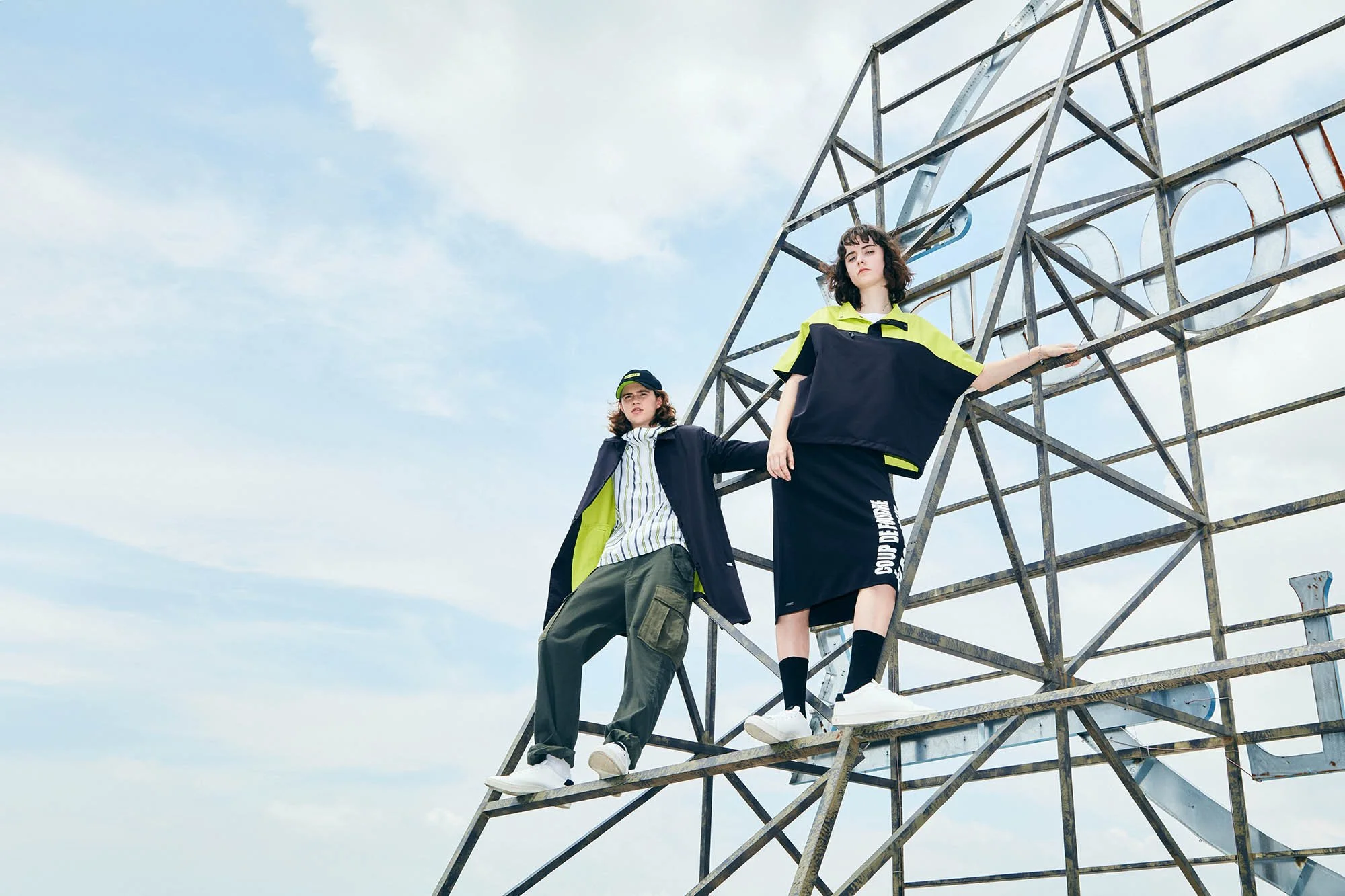 Two young women standing on a metal structure against a blue sky with clouds. One wears a baseball cap, a jacket, and cargo pants, while the other wears a color-block windbreaker, a skirt, and sneakers.