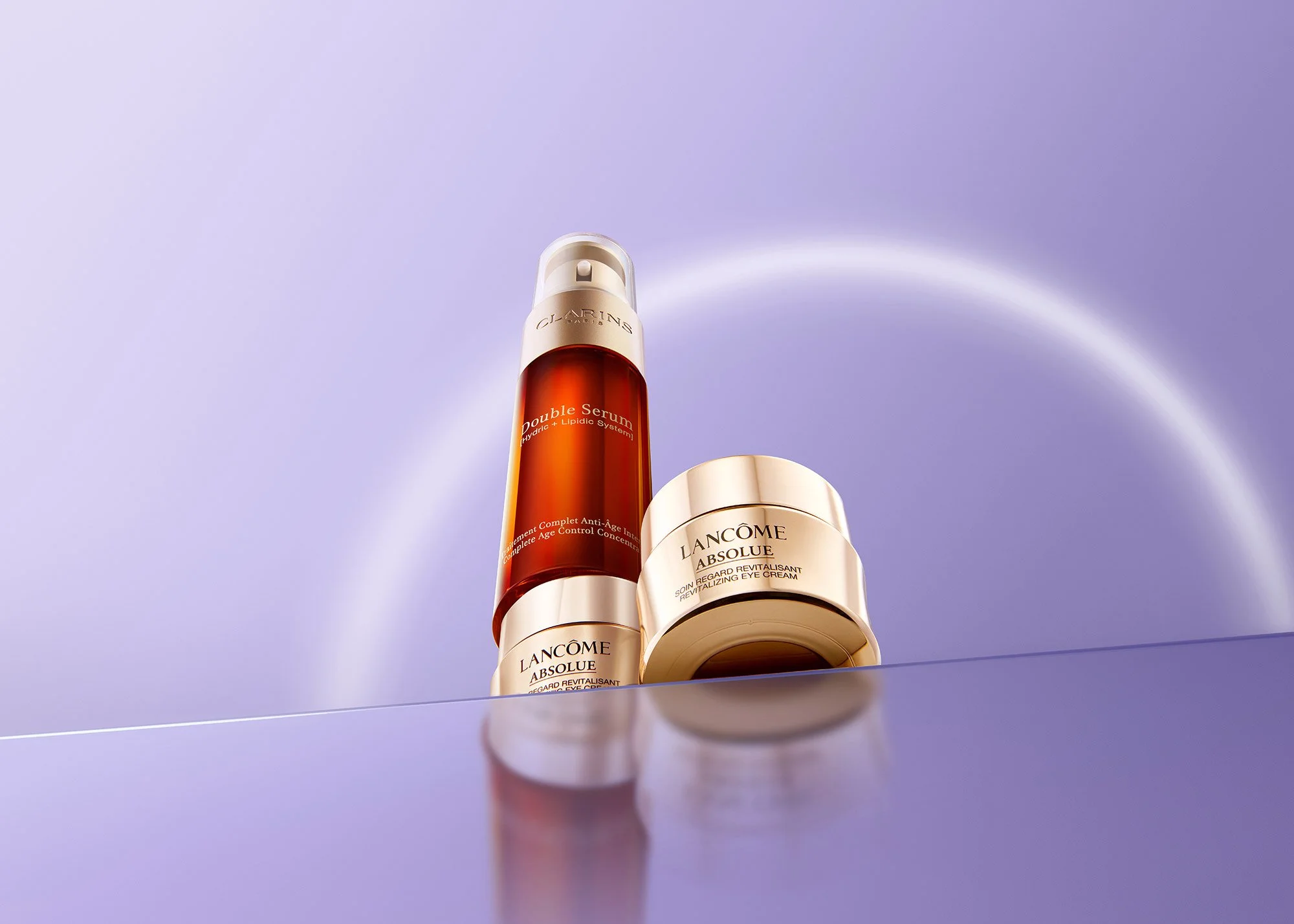 Two Lancôme skincare products, a serum and an eye cream, are displayed against a purple background with a soft glow and a rainbow-like reflection. The serum is in an orange bottle with a white pump, and the eye cream is in a gold jar.