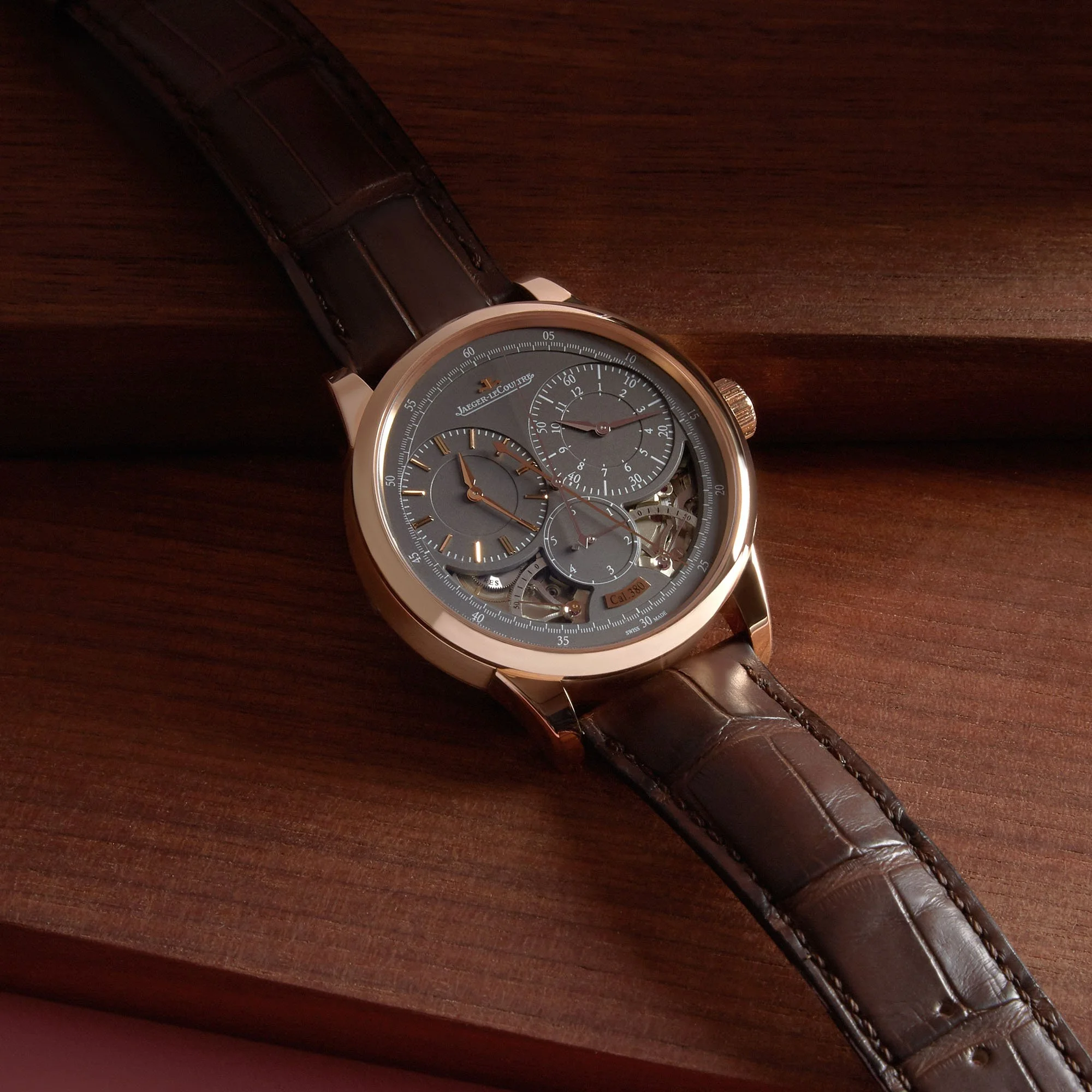 Close-up of a luxury wristwatch with a dark brown leather strap, silver case, and a complex gray dial showing multiple subdials and mechanical details, placed on a wooden surface.