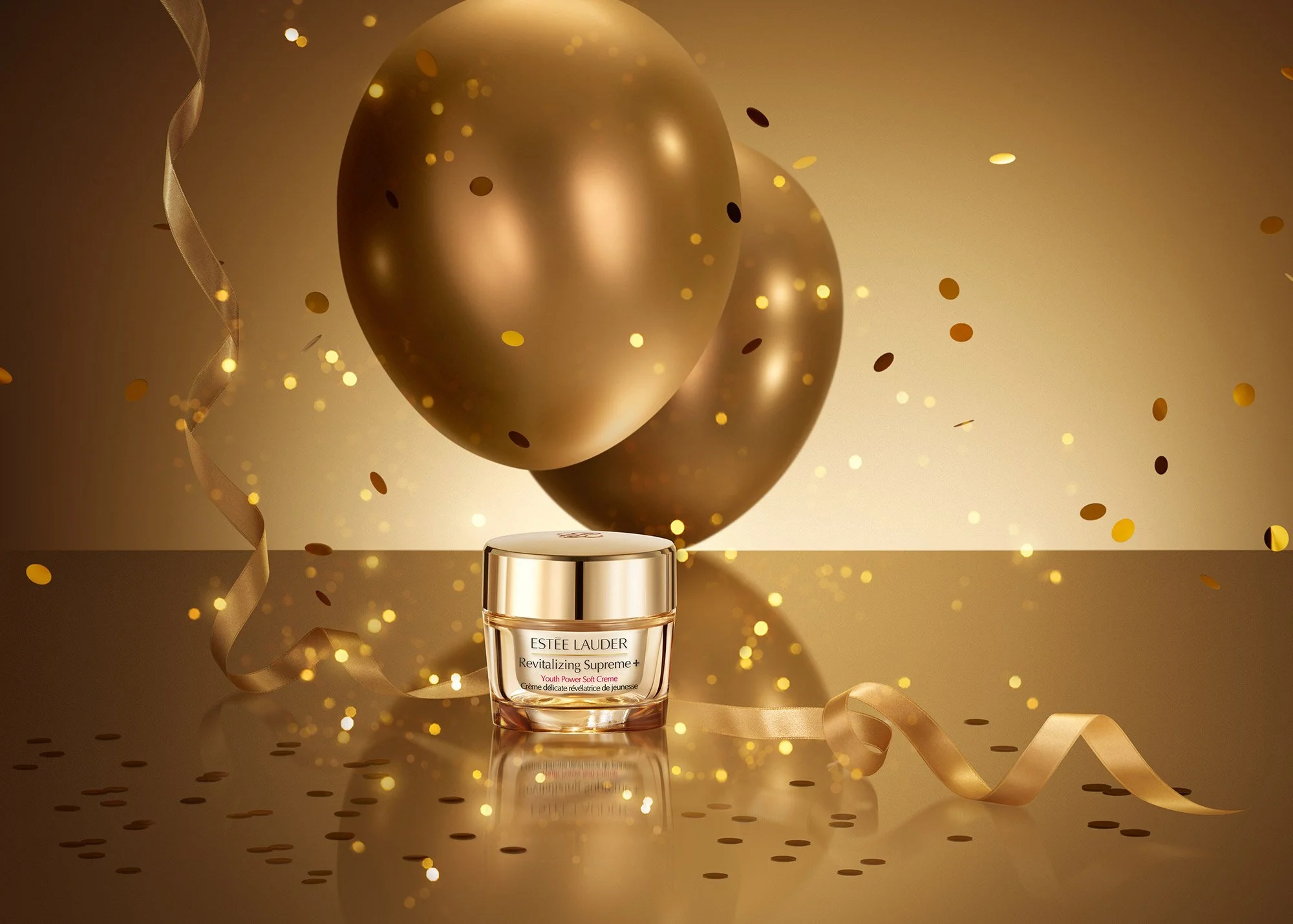 A jar of Estée Lauder Revitalizing Supreme+ Youth Power Soft Crème on a reflective surface, surrounded by gold balloons, gold confetti, and curled gold ribbons, celebrating a festive occasion.
