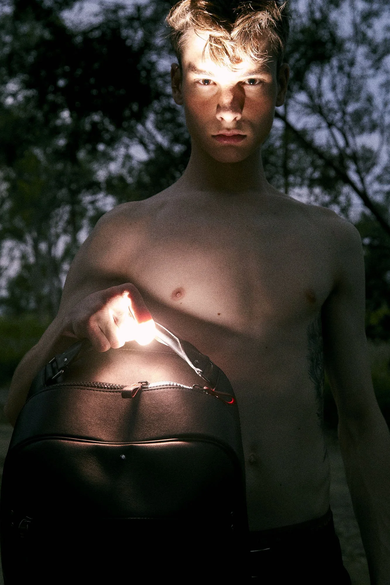 A shirtless young man holding a black backpack with a flashlight in a dark outdoor setting surrounded by trees.