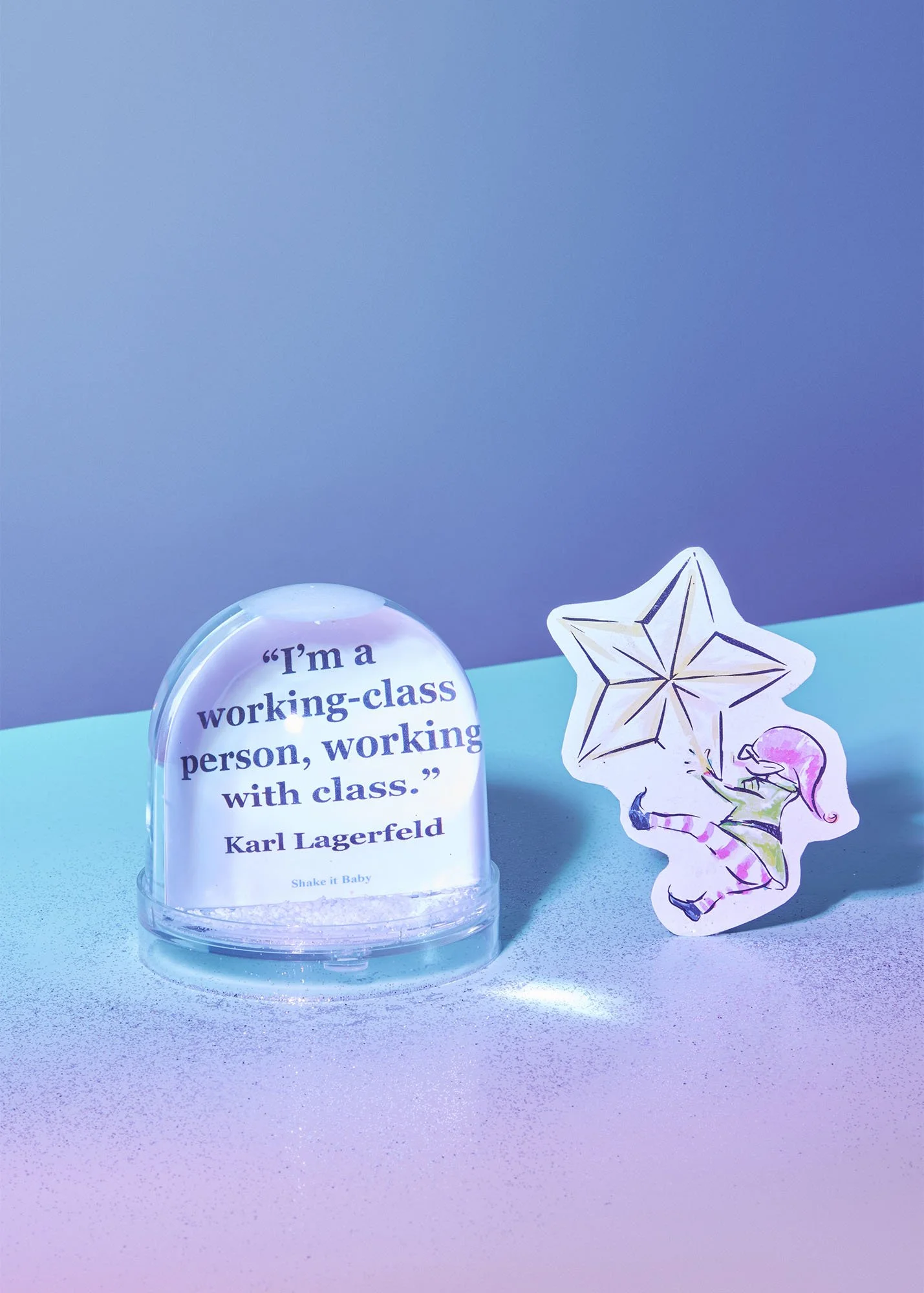 A snow globe with a quote by Karl Lagerfeld inside, reading 'I'm a working-class person, working with class.' next to a decorative sticker of a girl with pink hair holding a star-shaped kite.