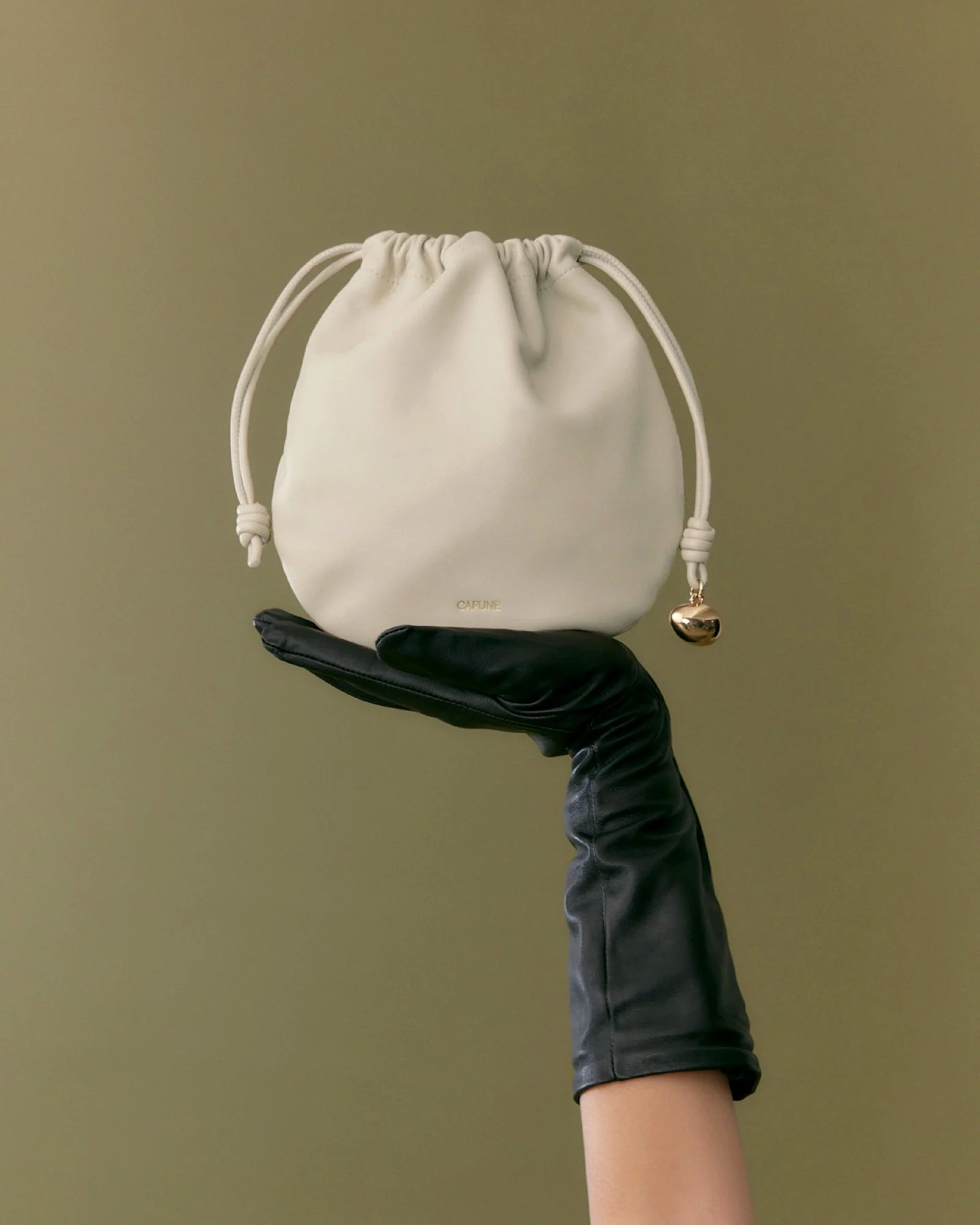 Person wearing black gloves holding a small white drawstring pouch. The pouch has the word "CAFUNE" printed on it and a metallic bell charm attached to the drawstring.