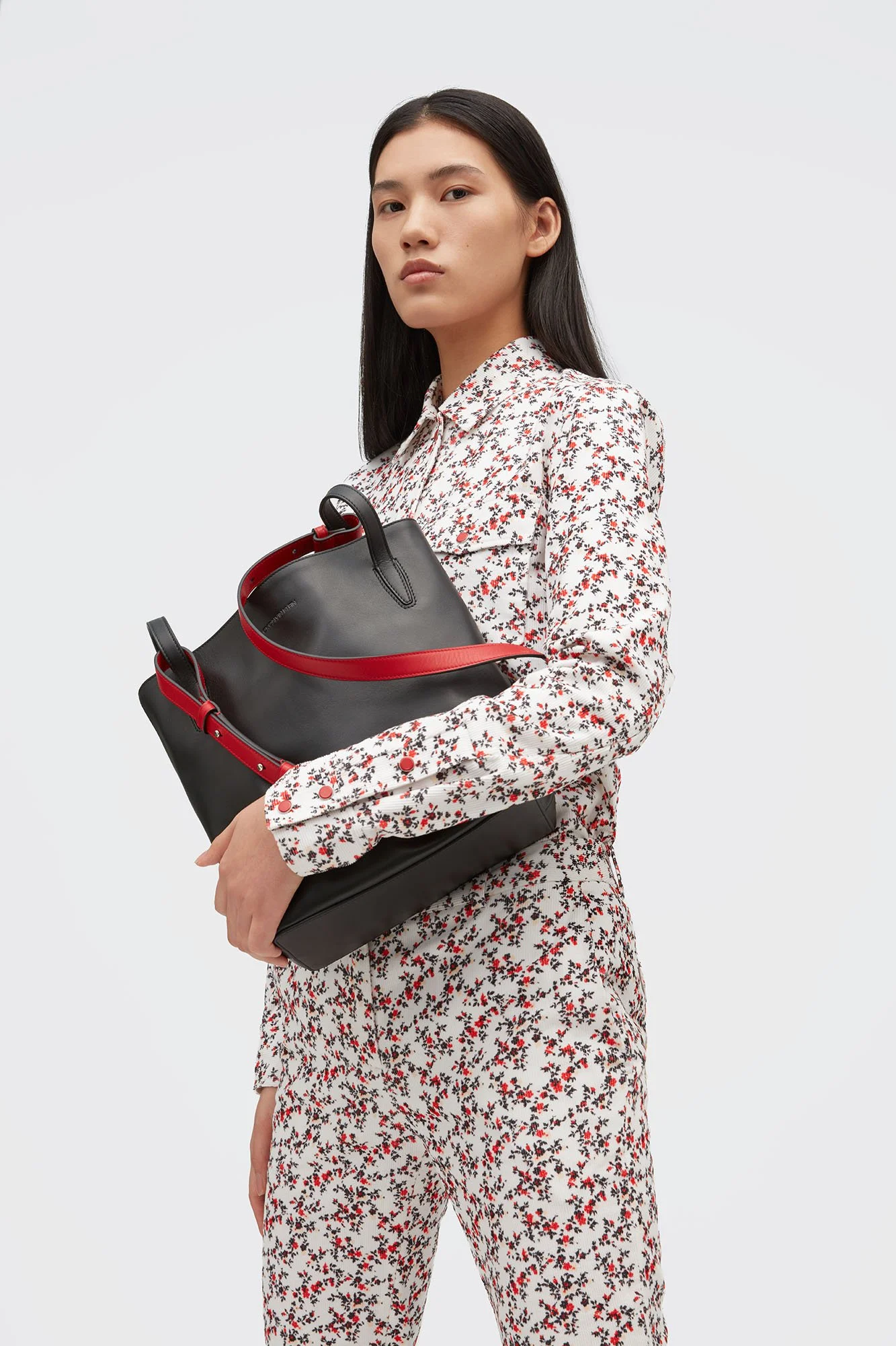 A woman with long dark hair wearing a white floral blouse and matching floral pants, holding a black handbag with red accents, posing against a plain light background.