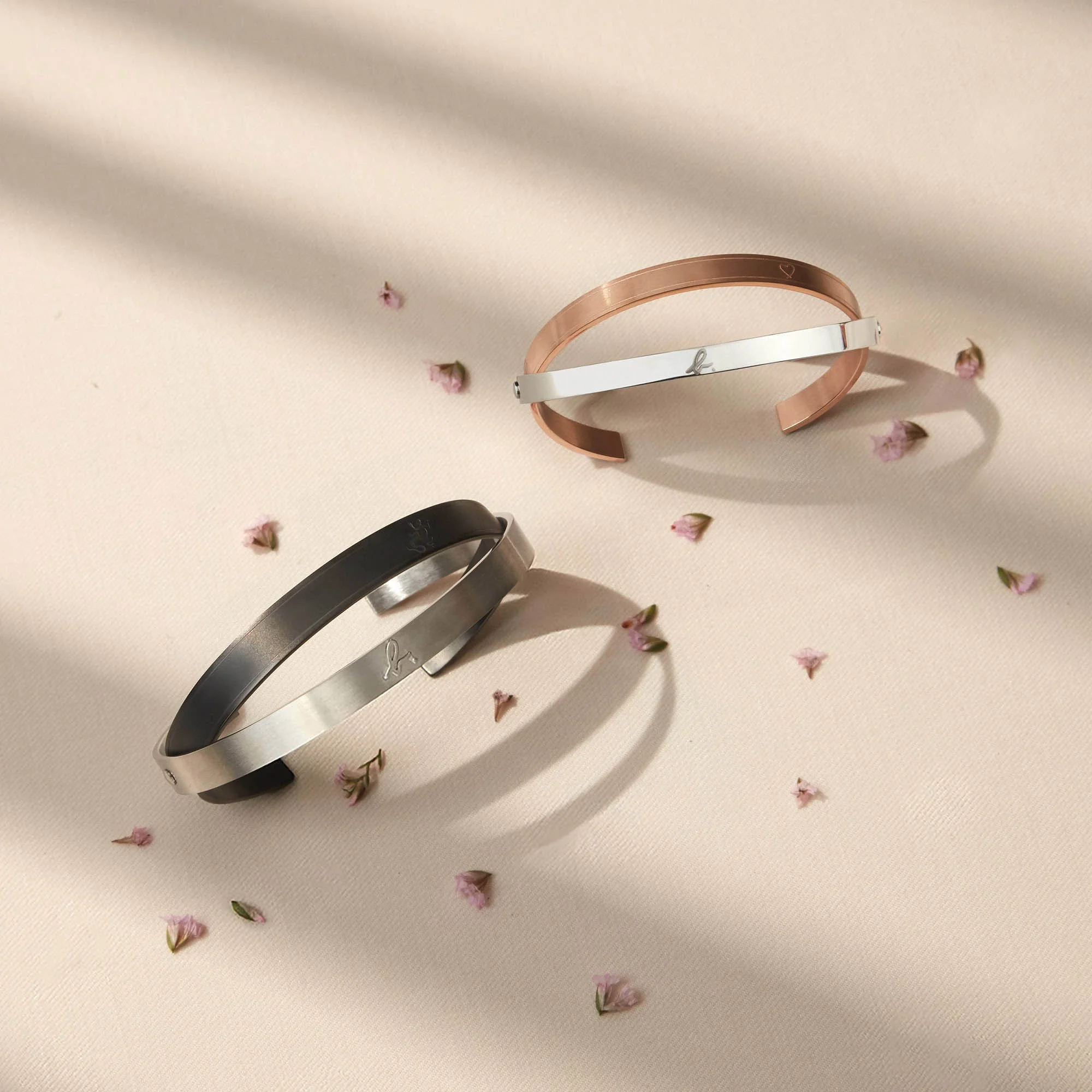 Three metal bracelets—black, silver, and rose gold—on a light fabric background with small pink flowers. The bracelets have a minimalist design with an engraved heart symbol.