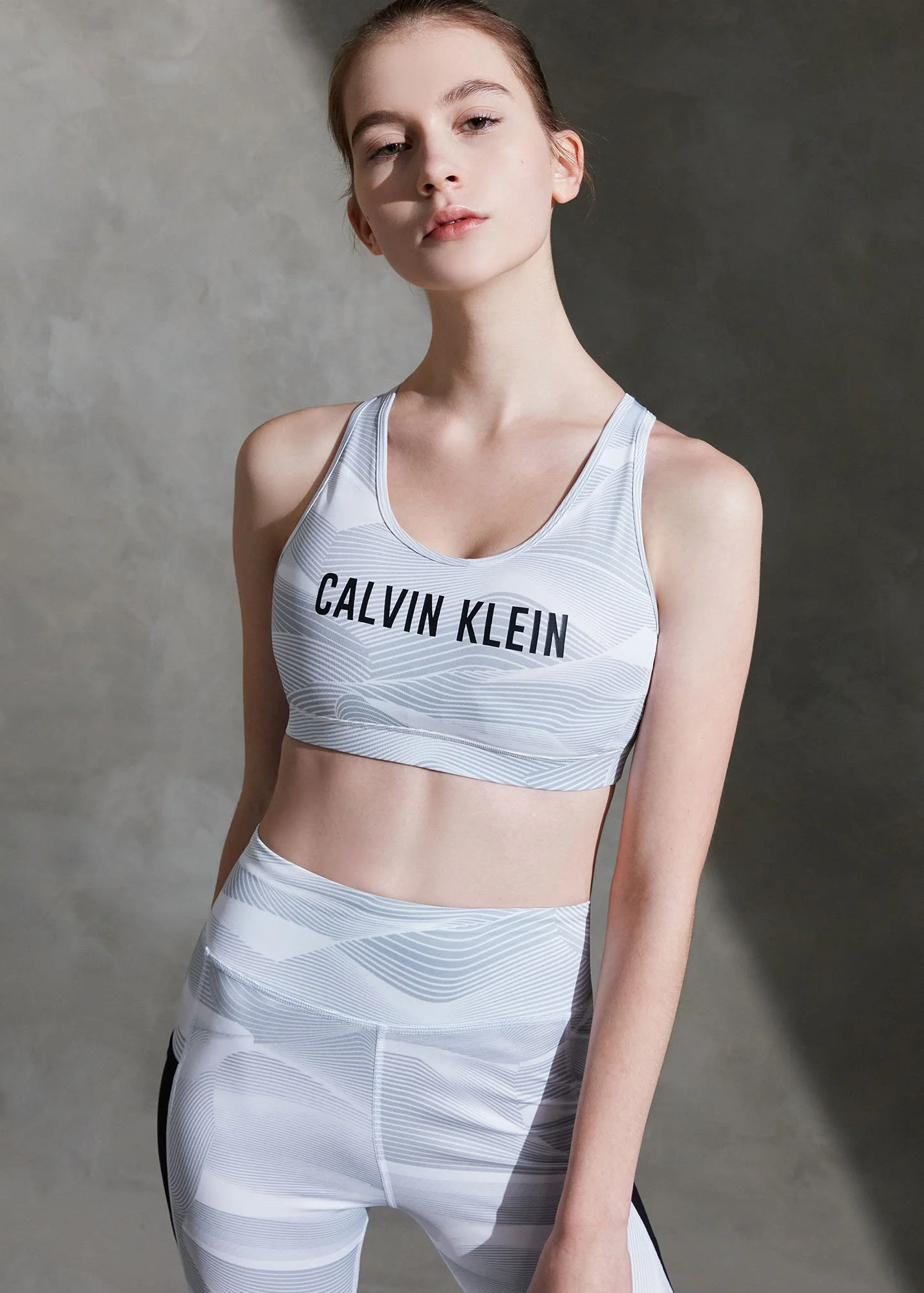 A young woman wearing a white Calvin Klein sports bra and matching leggings with a subtle pattern, standing against a neutral gray background.