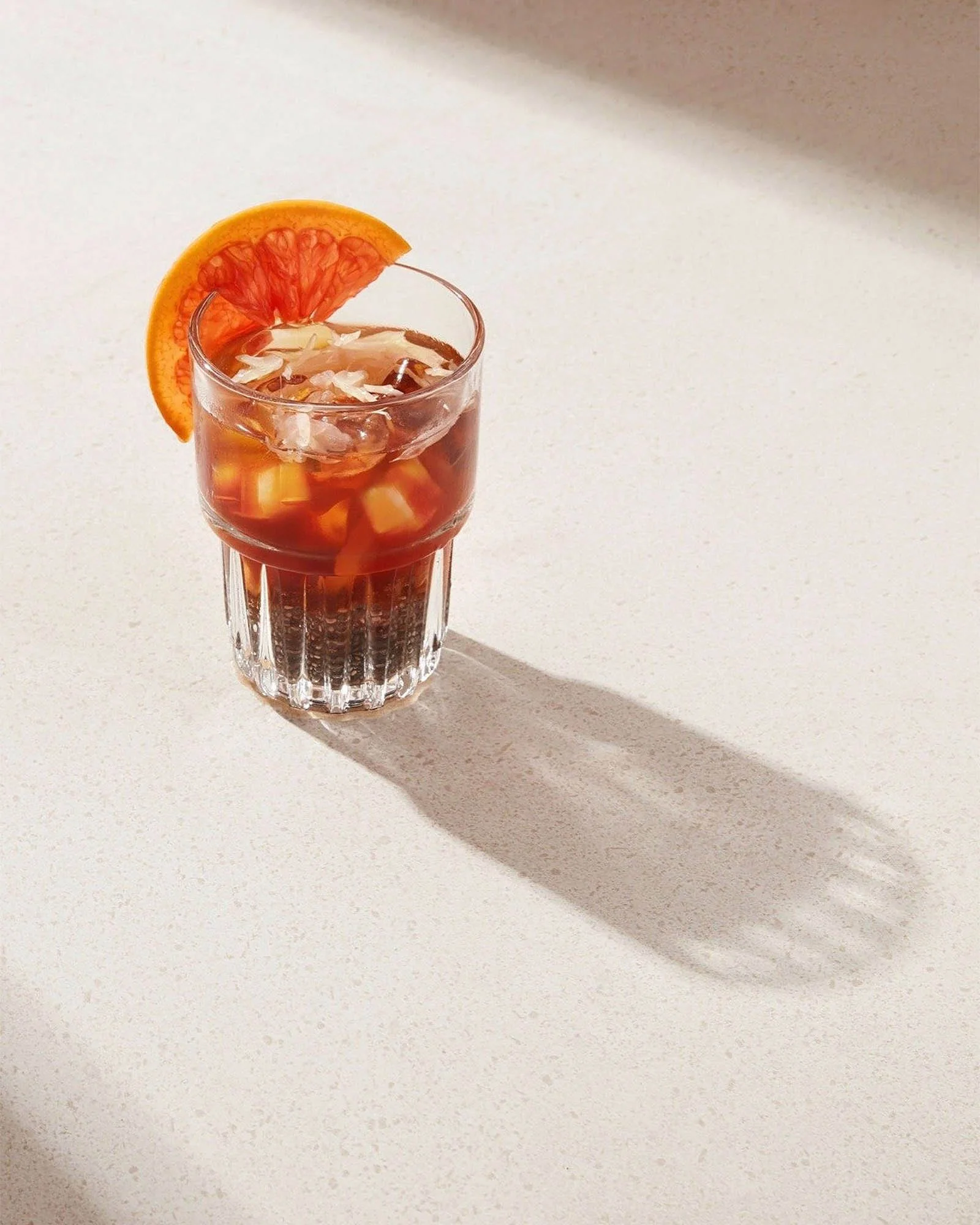 A glass of iced beverage garnished with a slice of blood orange, casting a long shadow on a white surface.