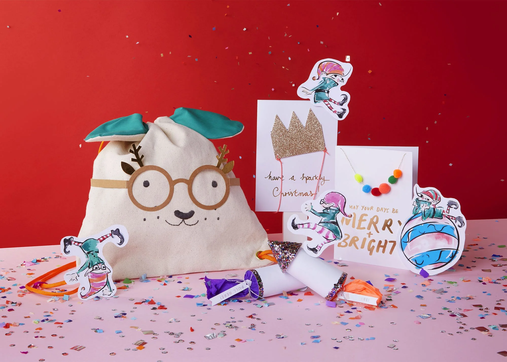 Decorative Christmas scene with a plush reindeer bag wearing glasses, surrounded by cut-out cartoon characters, colorful confetti, and handmade cards with festive messages on a pink and red background.