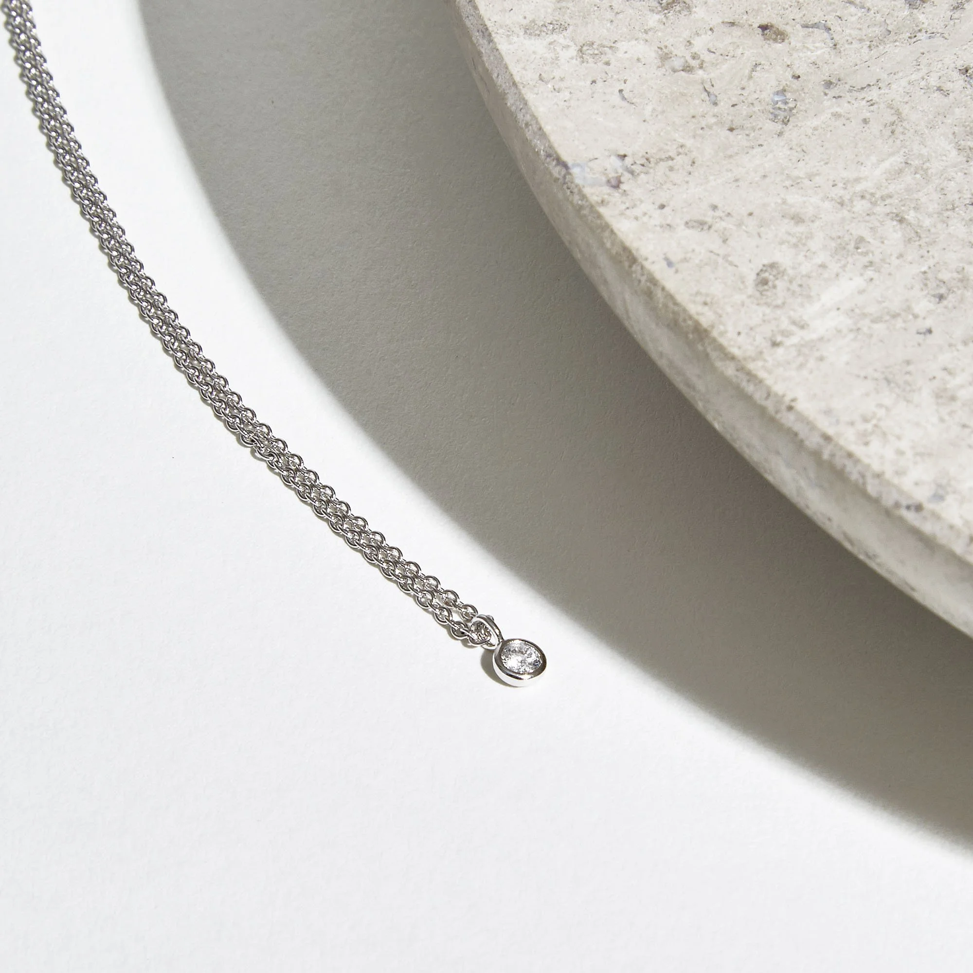 A silver necklace with a circular pendant containing a single small diamond, resting on a white surface near a piece of beige stone.
